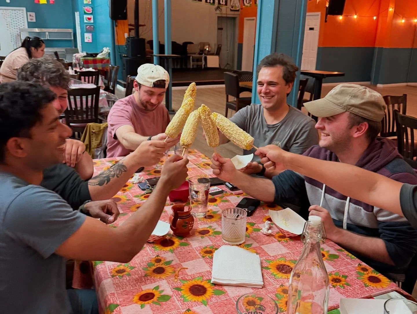 Group of friends sitting around a table, smiling, holding and toasting with corn cobs on sticks in a restaurant with colorful decor.