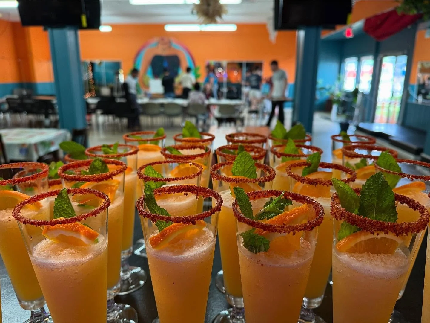 Multiple glasses of orange-colored drinks garnished with orange slices, mint leaves, and chili powder rimmed glasses on a table in a colorful restaurant or cafe.
