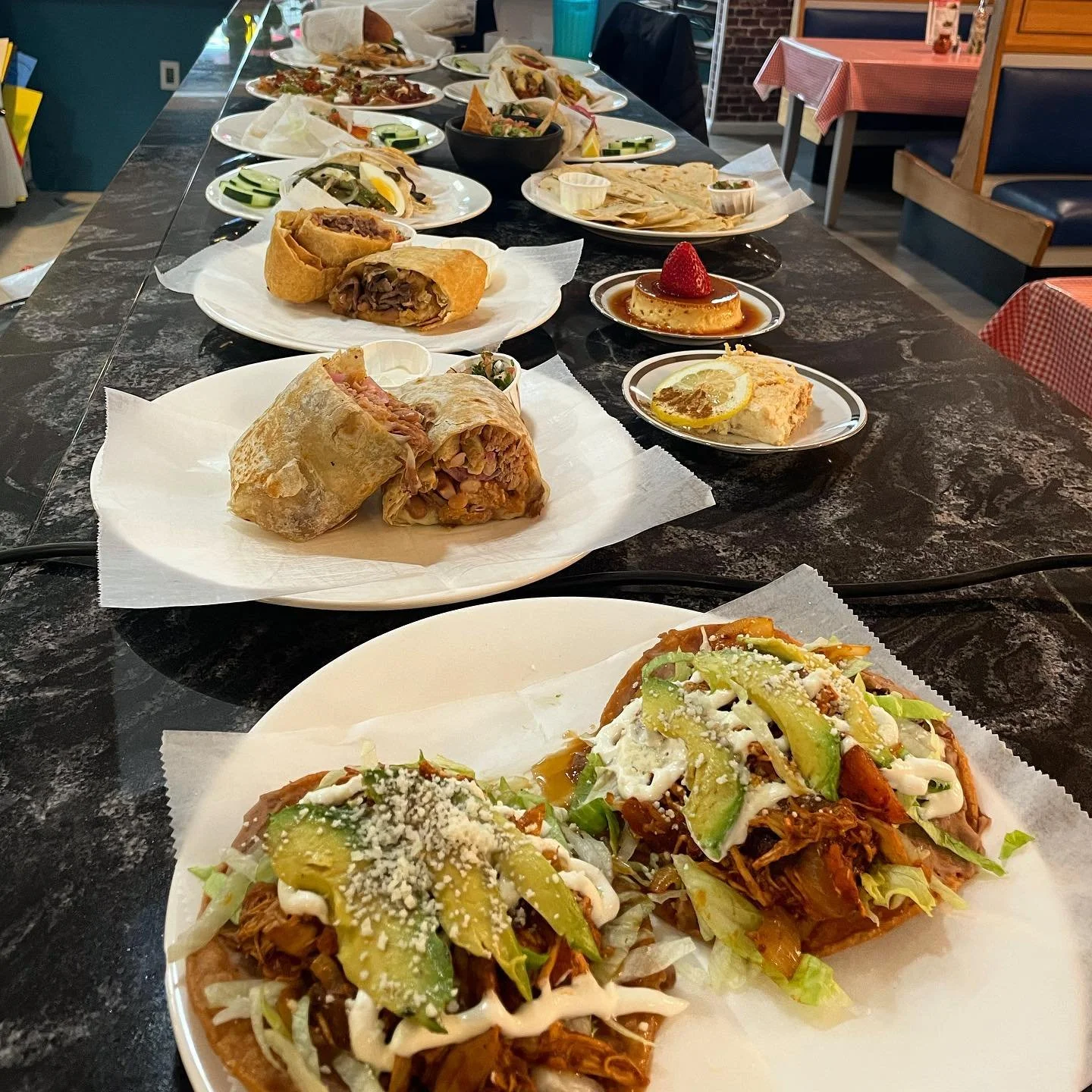 A long black table filled with plates of Mexican food, including tacos, burritos, quesadillas, and small dessert plates with flan, cheesecake, and pudding, in a restaurant setting.