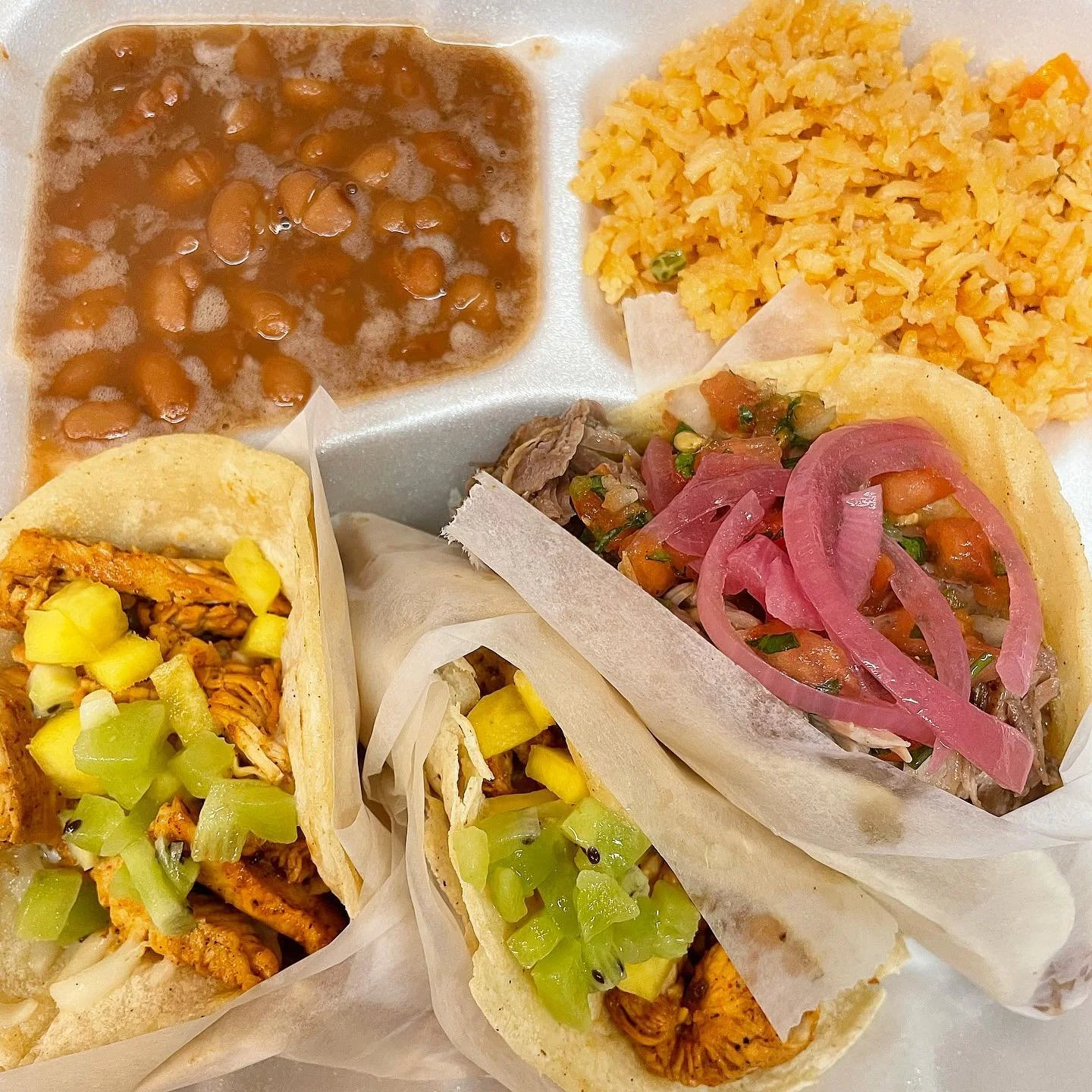 A plate of Mexican food including two tacos filled with shredded chicken, chopped celery, and yellow peppers, a side of Mexican rice, and beans with sauce.