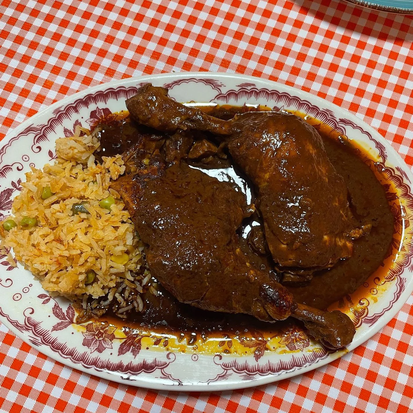 Plate with cooked chicken covered in dark sauce, rice mixed with vegetables, and cabbage, on a checkered red and white tablecloth.