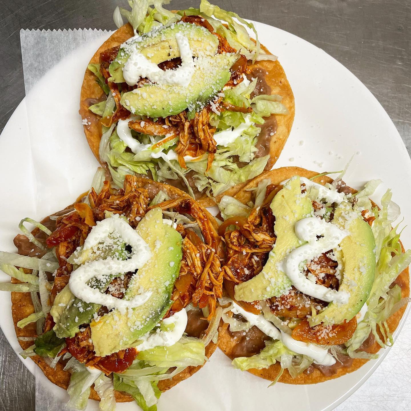 Three tacos on a white plate topped with shredded lettuce, avocado slices, shredded meat, sour cream, and grated cheese.