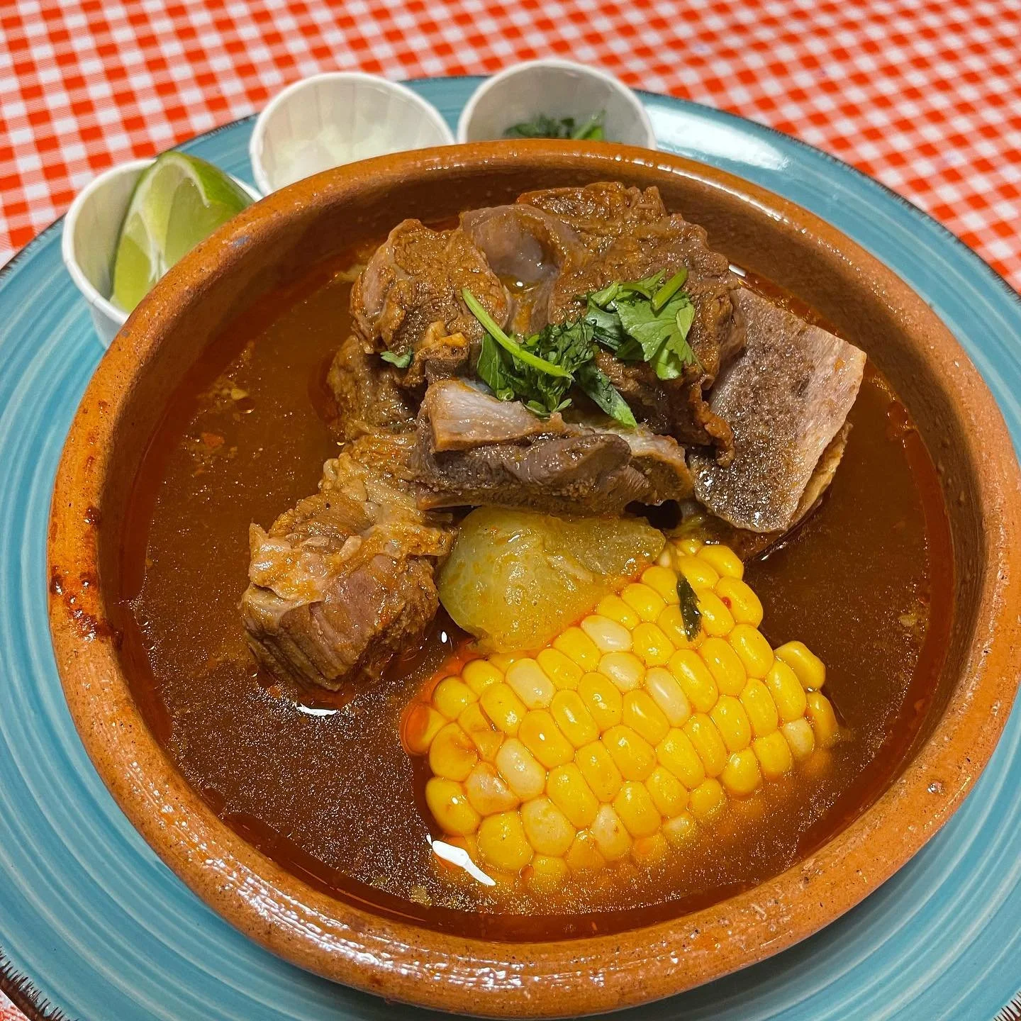A bowl of Mexican beef stew with corn, potatoes, chilis, topped with cilantro, served with lime and condiments