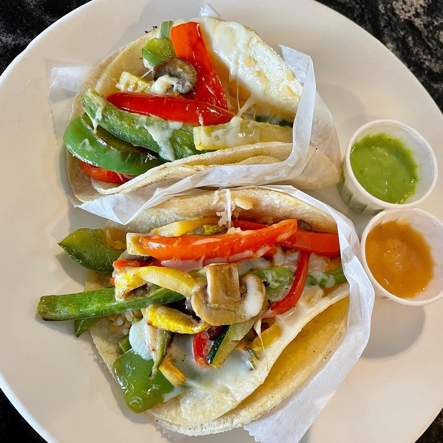 Two open-faced vegetable tacos with red, green, and yellow peppers, mushrooms, on soft tortillas, served with green and orange sauces.