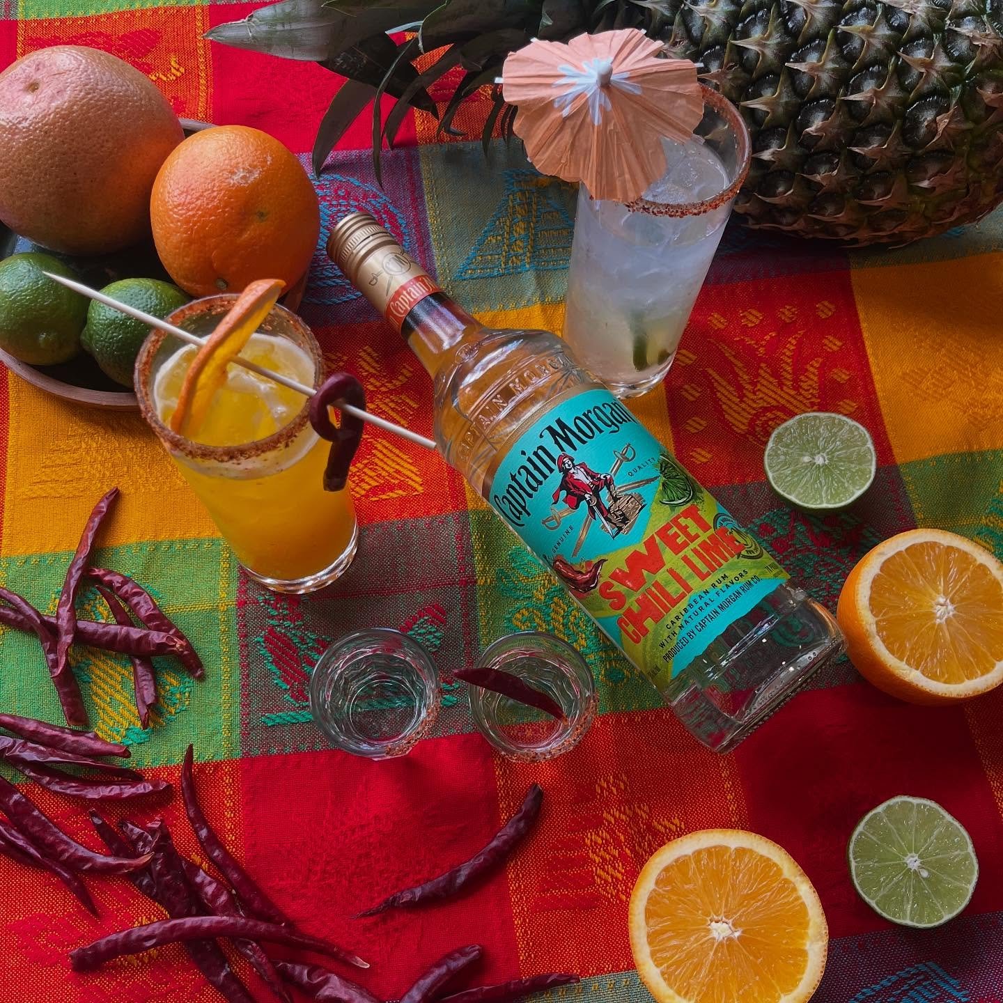 A colorful table setting with tropical fruits, including pineapples, oranges, limes, and dried red chili peppers. A bottle of Captain Morgan Sweat Chili Lime flavored rum is placed among two prepared cocktails with garnishes of citrus slices and umbrellas.