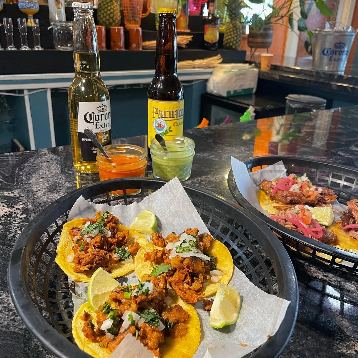 Two baskets of street-style tacos on a black marble counter, garnished with lime wedges and chopped onions, with bottles of beer and small containers of hot sauce and guacamole in the background at a Mexican restaurant.