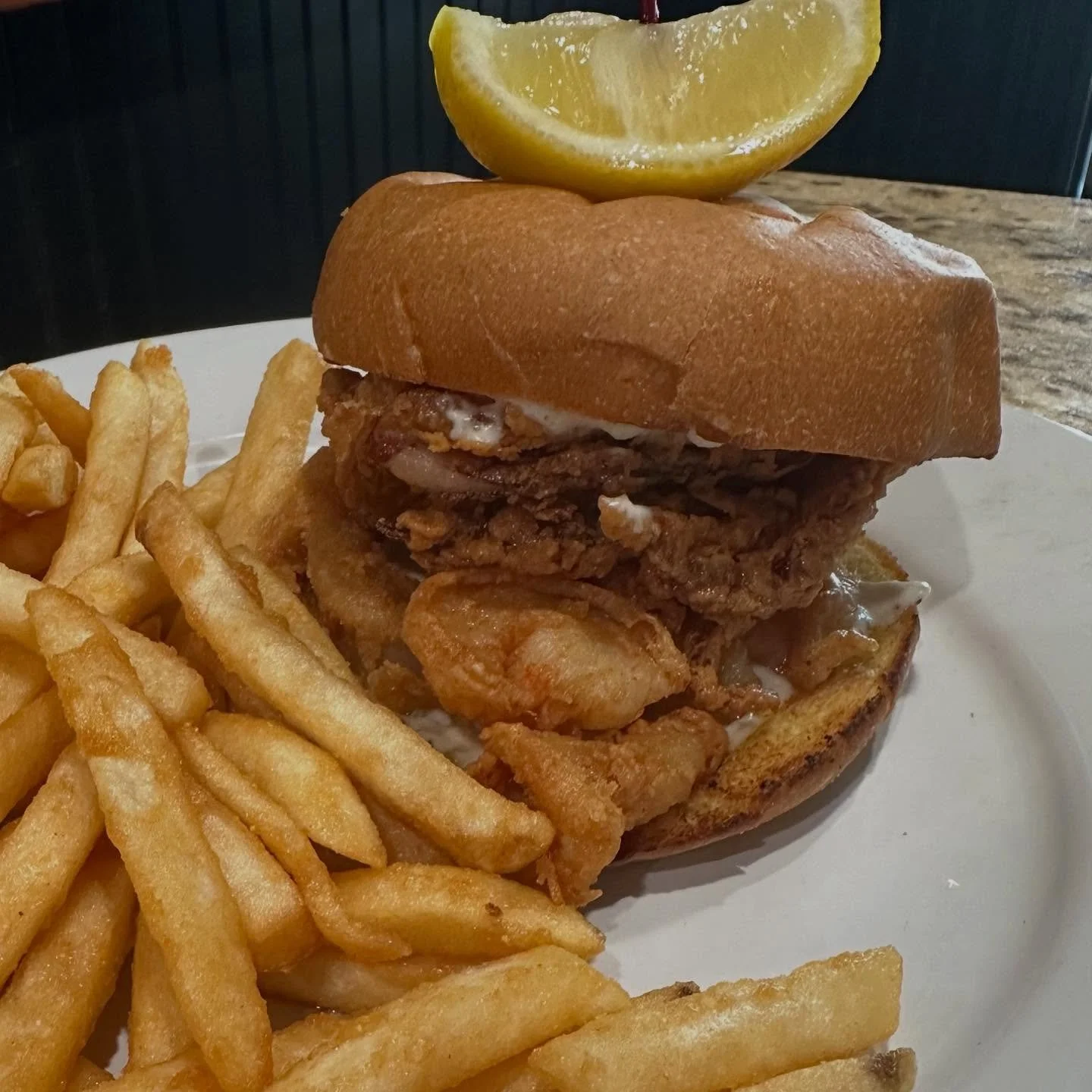 A plate with a fried chicken sandwich topped with a lemon wedge, served with French fries on the side.