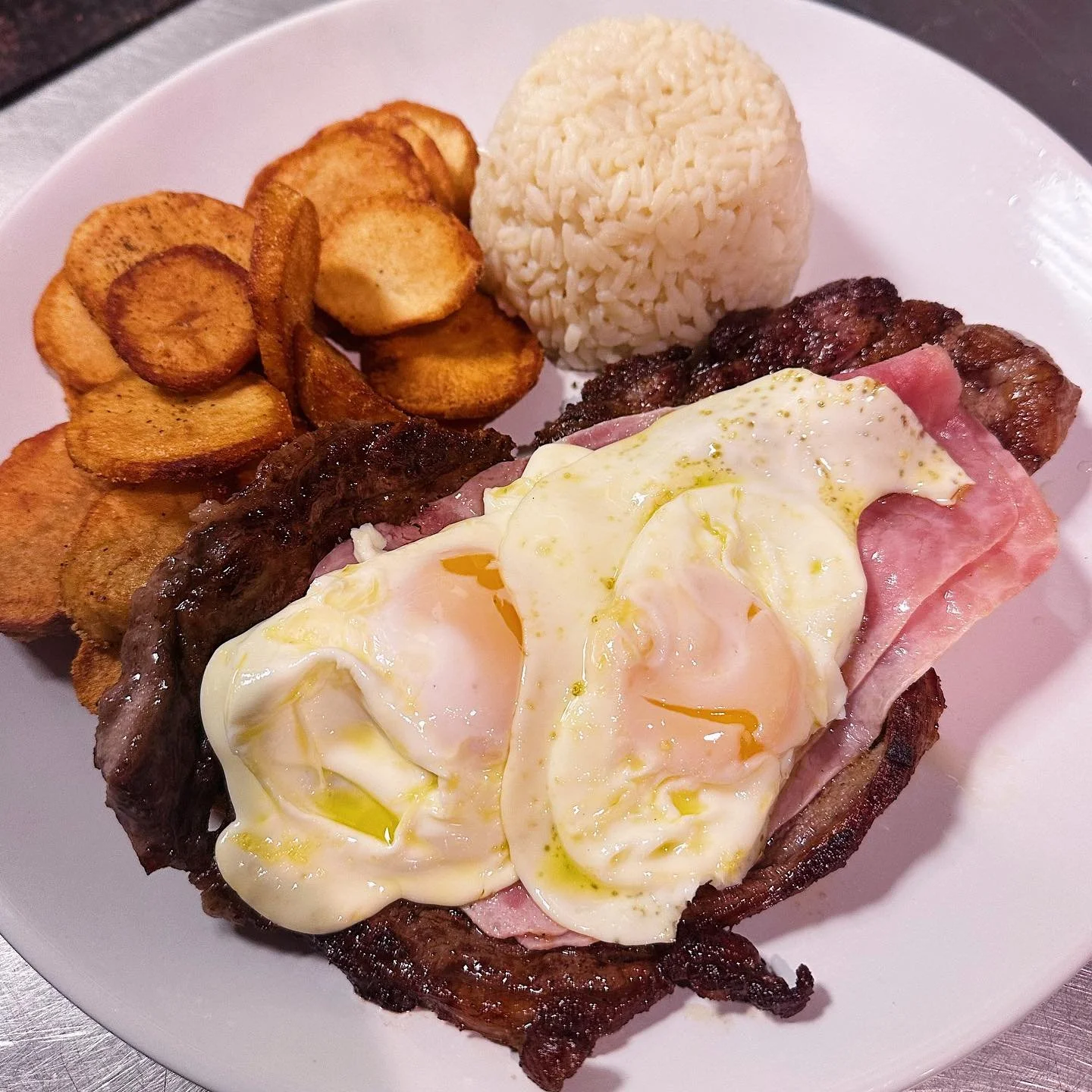 Plate of food with fried potatoes, rice, grilled meat, ham, bacon, and two fried eggs.