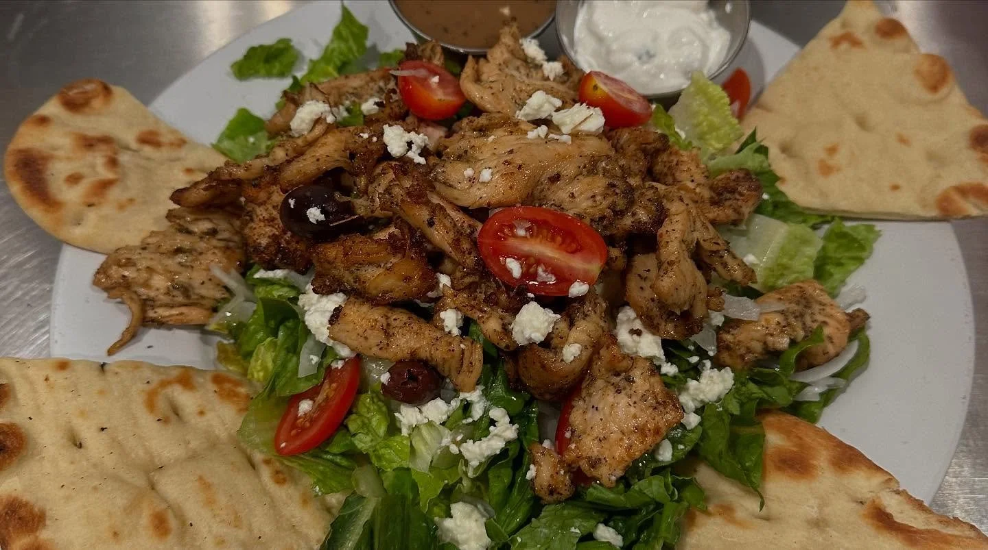 A plate of grilled chicken atop a bed of lettuce, cherry tomatoes, feta cheese, and black olives, with warm pita bread on the side and small bowls of white sauces in the background.