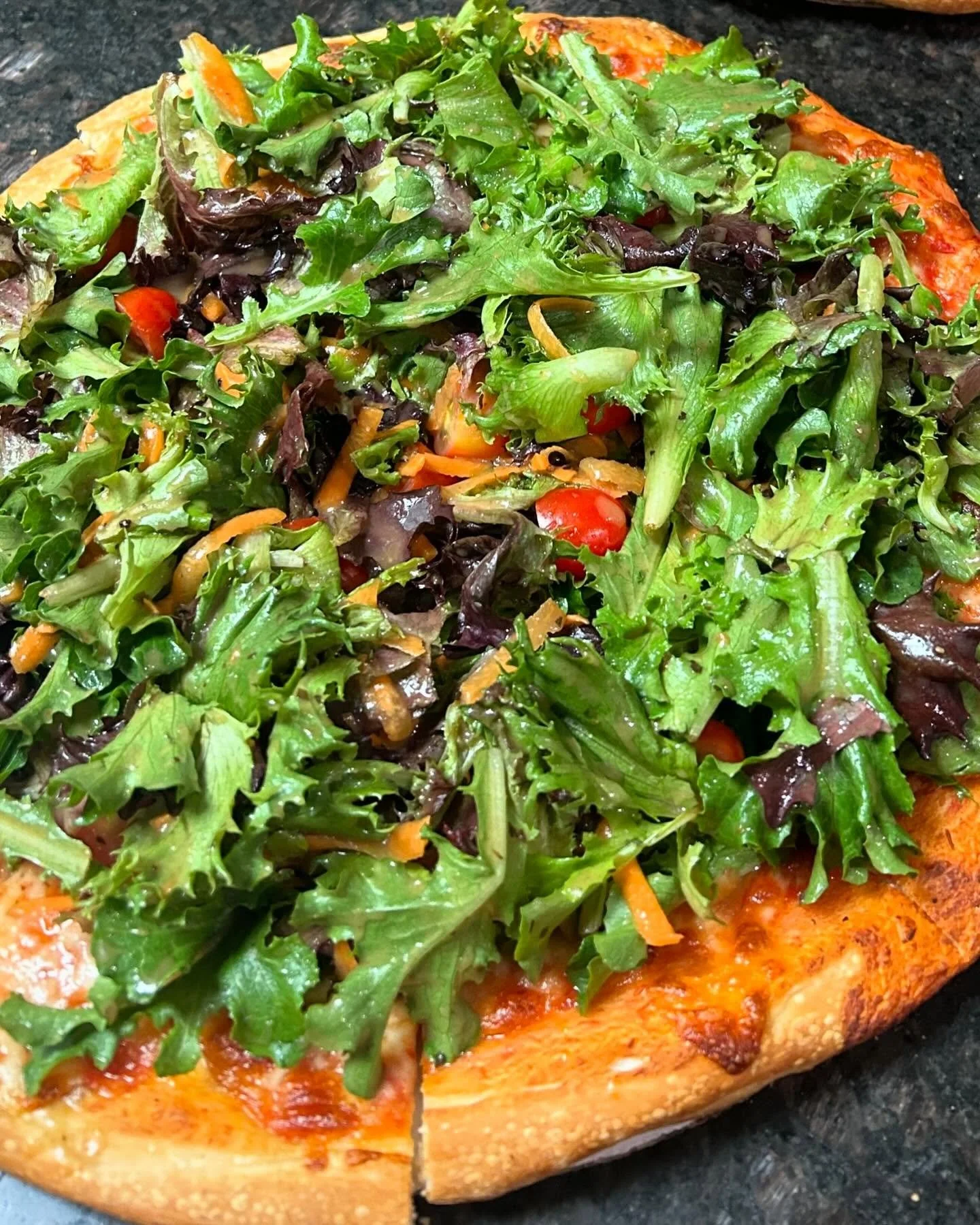 A pizza topped with a generous amount of mixed leafy greens, cherry tomatoes, shredded carrots, and a crispy crust.