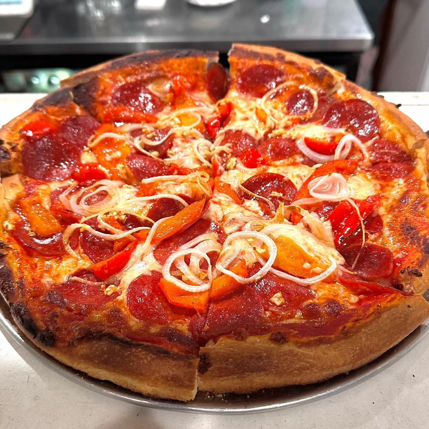 A freshly baked pepperoni pizza with slices of pepperoni, red and yellow bell peppers, and onion slices on a round metal tray.