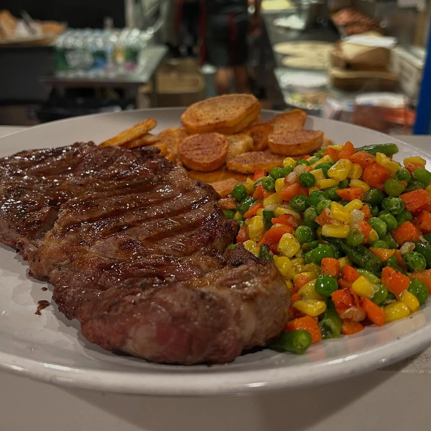 Plate of grilled steak, sautéed mixed vegetables including peas, corn, carrots, and green beans, sliced toasted bread, and potato chips in a restaurant setting.