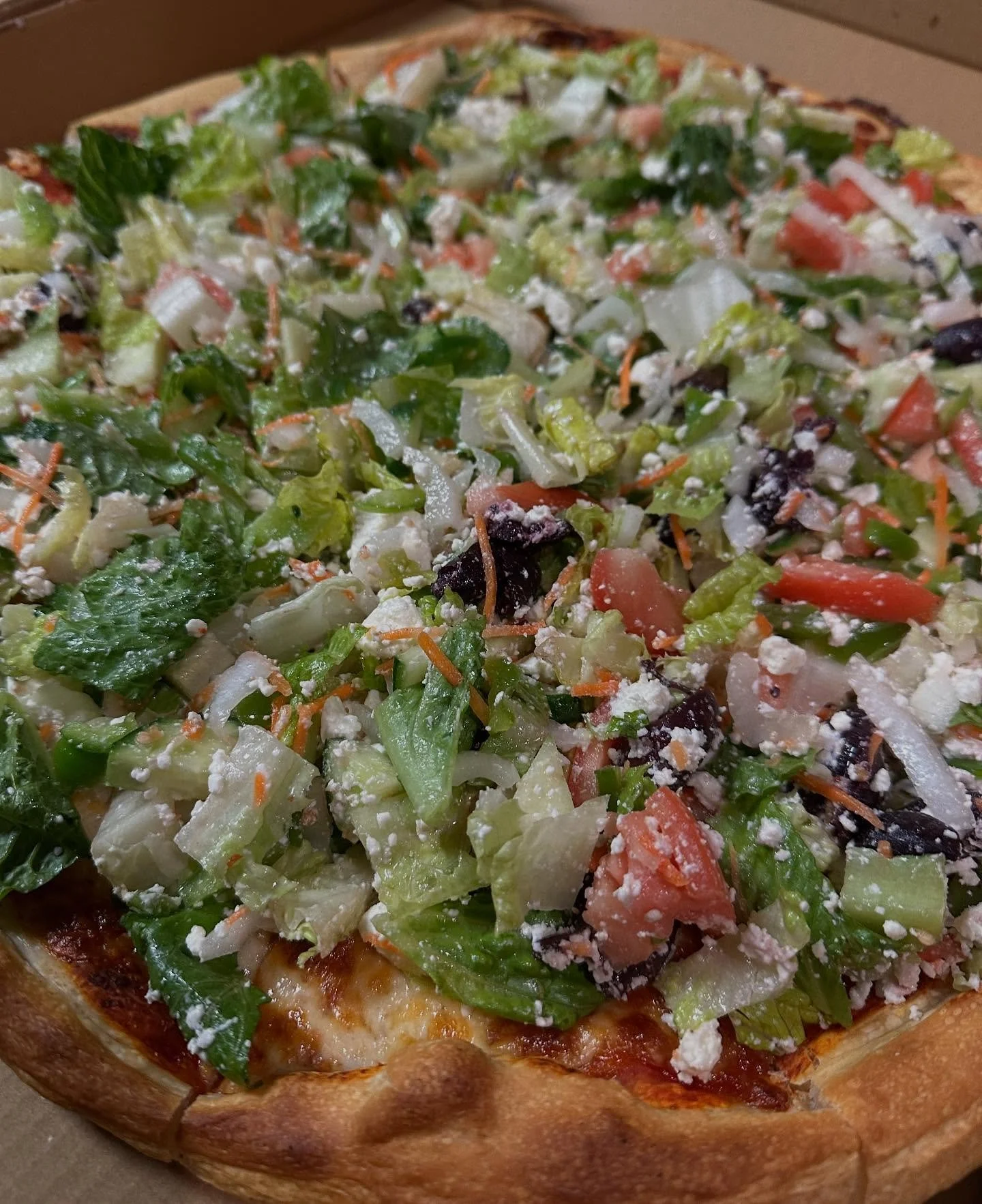Close-up of a pizza topped with lettuce, tomatoes, black olives, shredded carrots, and crumbled cheese.