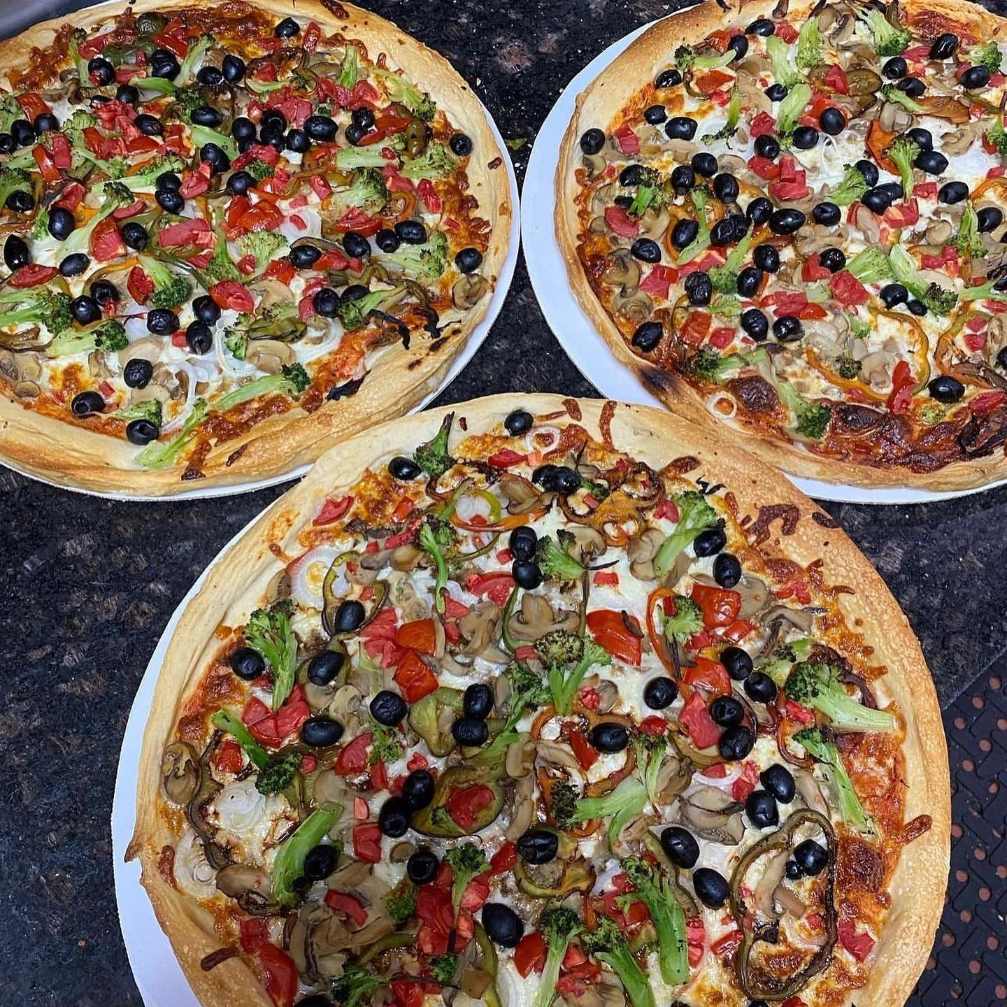 Three vegetable pizzas topped with black olives, red peppers, green broccoli, mushrooms, and onions on a dark countertop.
