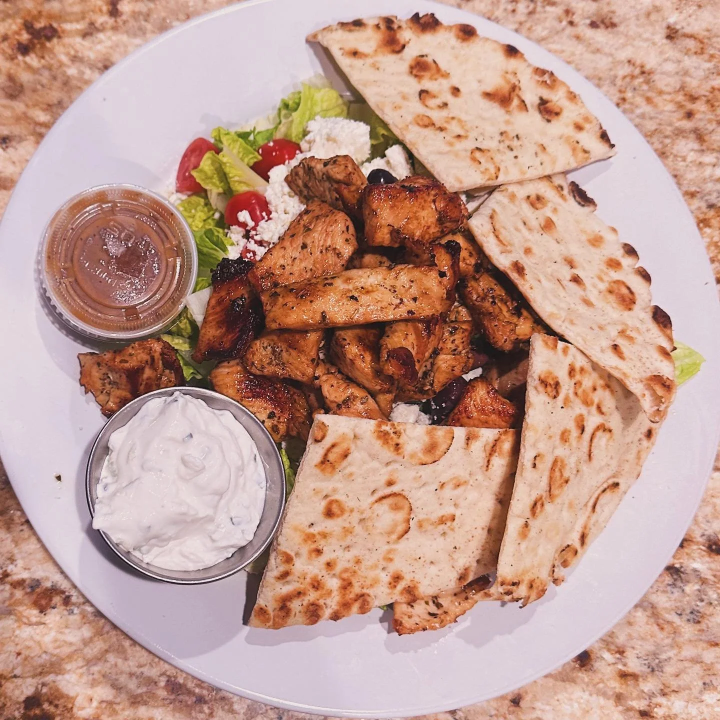A white plate of grilled chicken, flatbread, and salad with cherry tomatoes, cucumbers, lettuce, and feta cheese, with small containers of sauce and yogurt.
