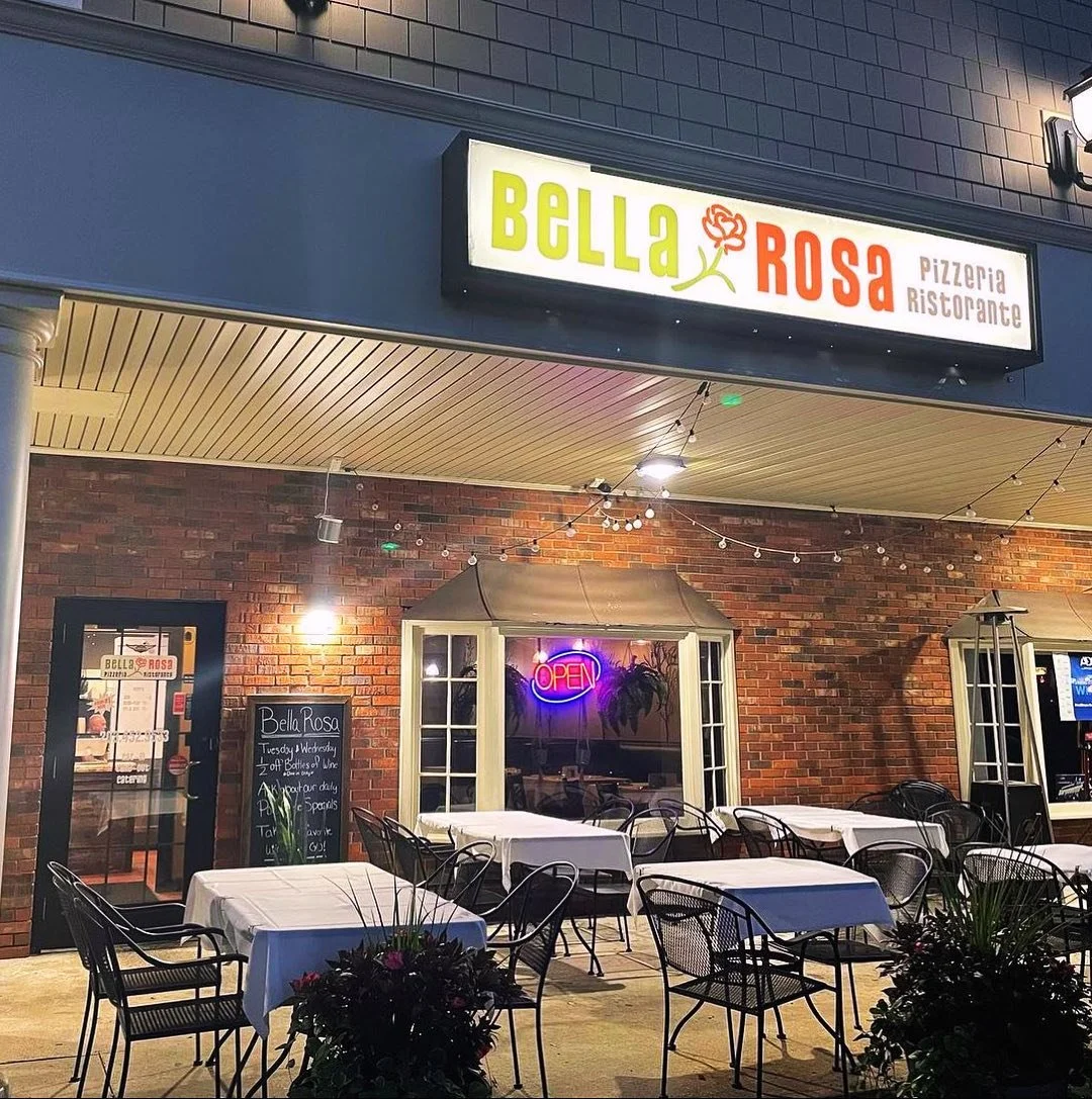 Outside view of Bella Rosa Pizzeria Ristorante with tables and chairs, neon 'OPEN' sign, brick wall, string lights, and greenery.