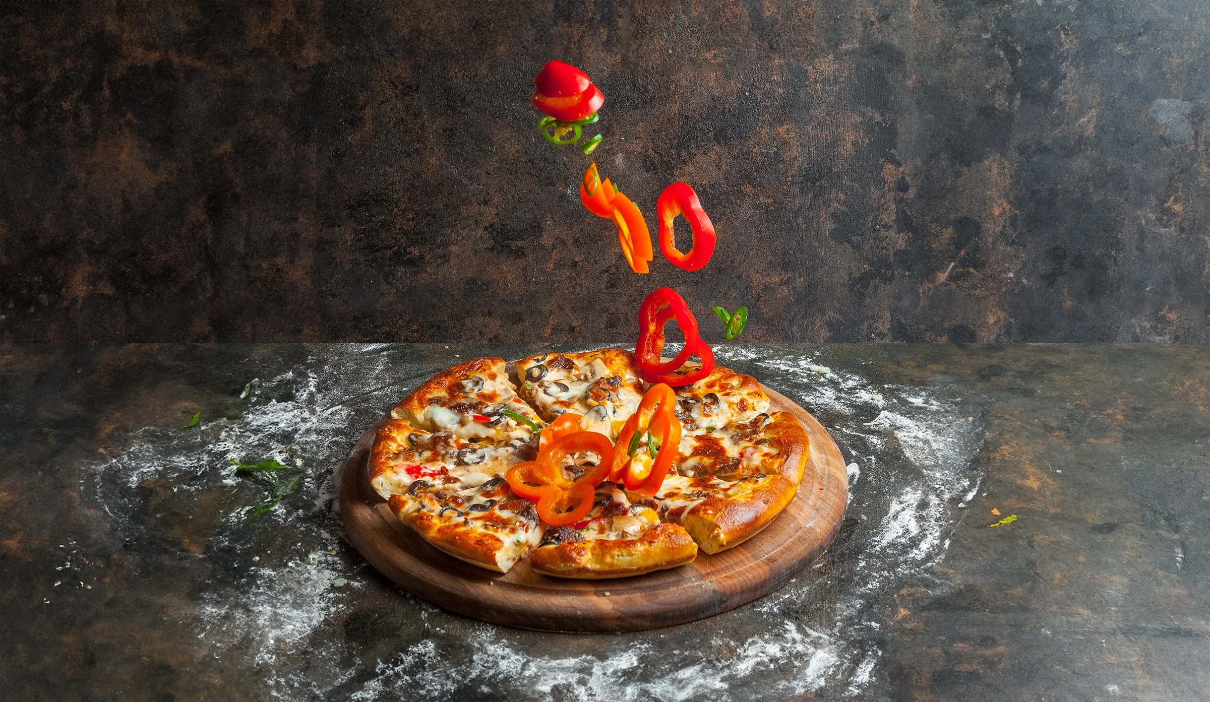 Sliced pizza on a round wooden board with sliced red and orange bell peppers falling onto it, with a flour-dusted dark surface background