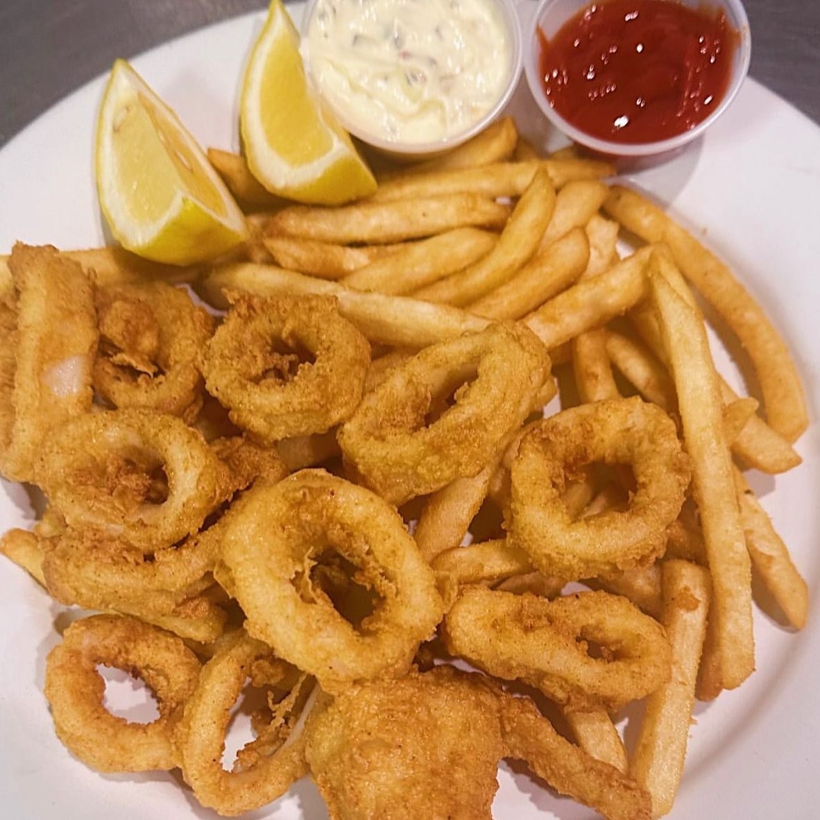 Plate of fried calamari rings with French fries, lemon wedges, and two dipping sauces, one tartar sauce and one ketchup.