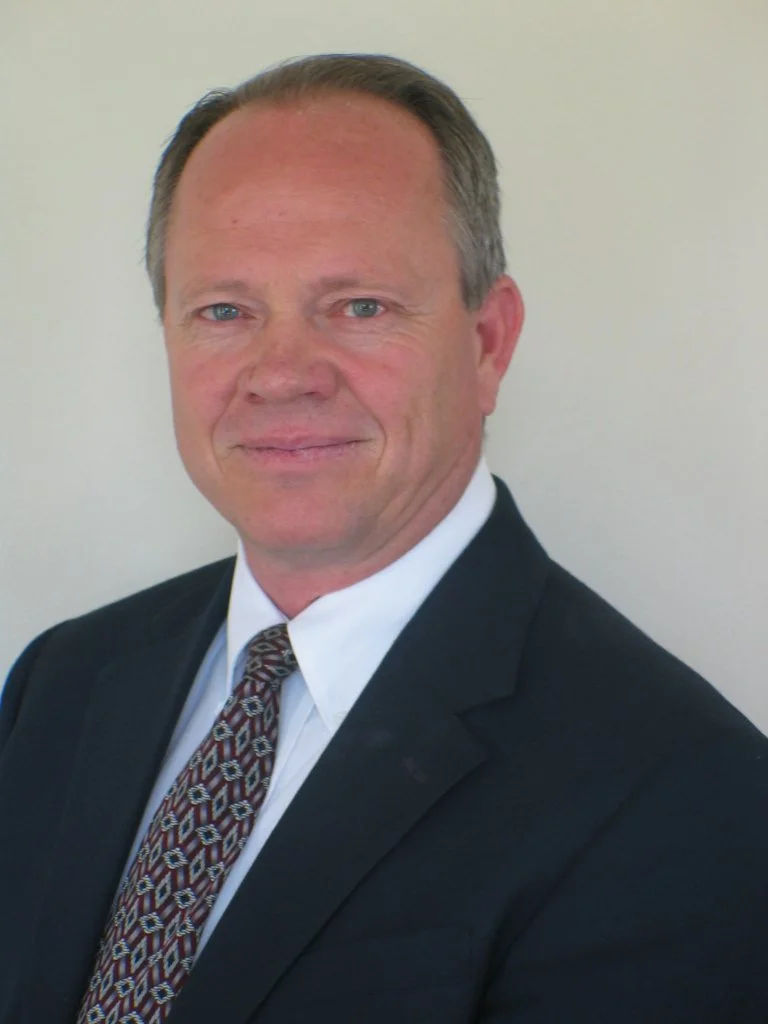 A middle-aged man in a dark suit, white shirt, and patterned tie, posing against a plain light-colored background.