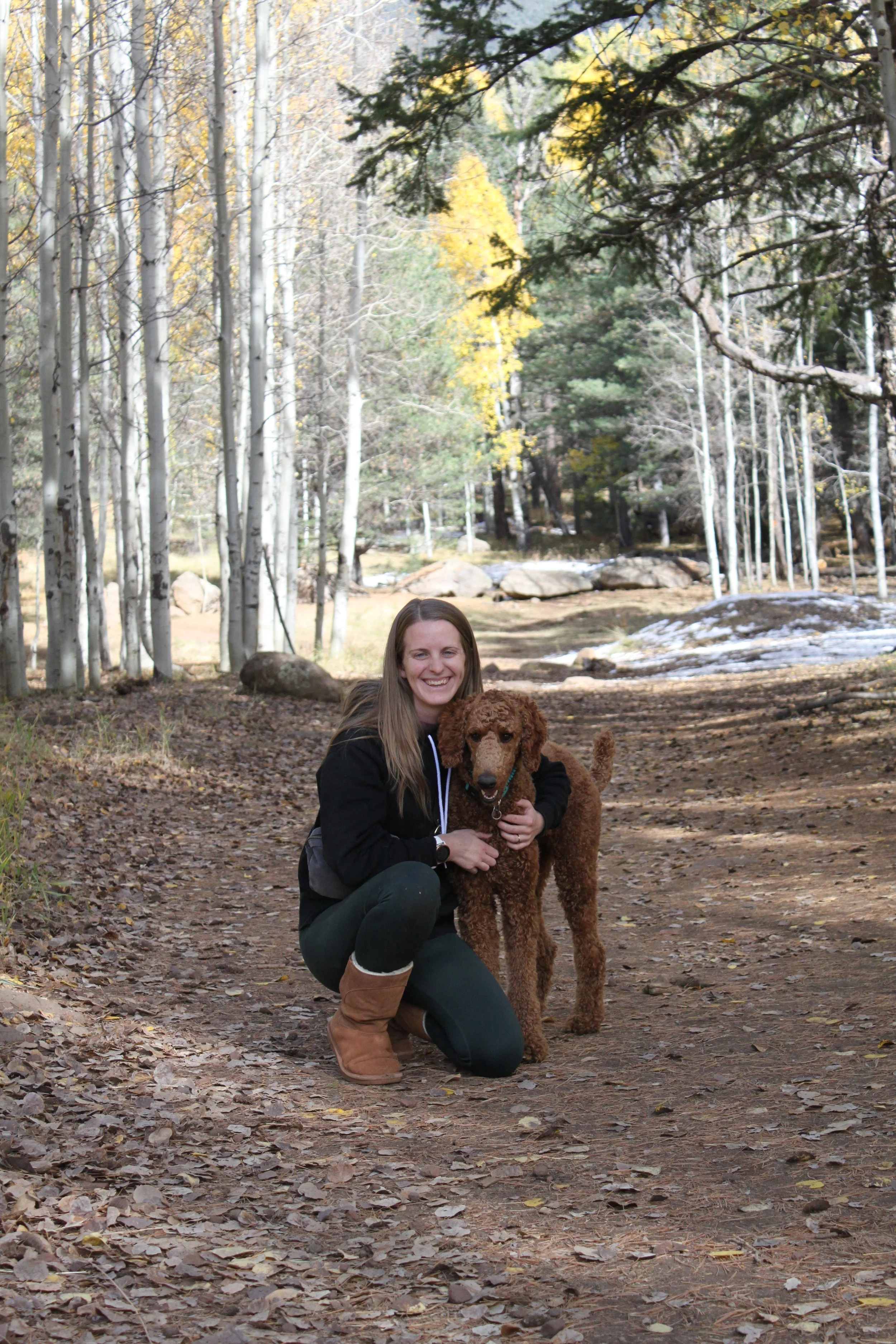 A woman in outdoor clothing kneeling on a forest trail, smiling, with a brown poodle dog.