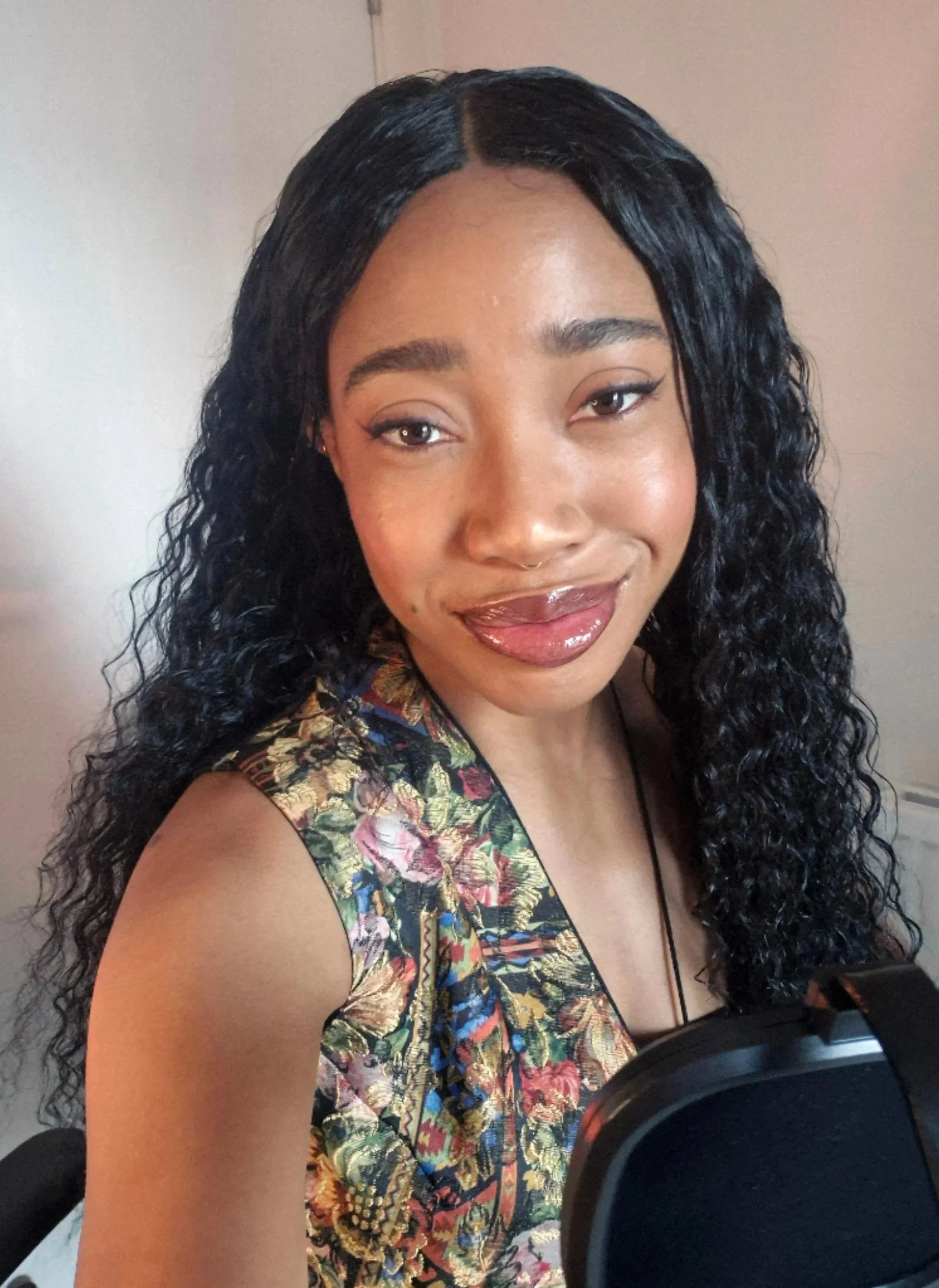 Kiara Zhanae. A young woman with long, curly black hair, wearing glossy lipstick and a colourful floral sleeveless vest, smiling at the camera with a microphone below.