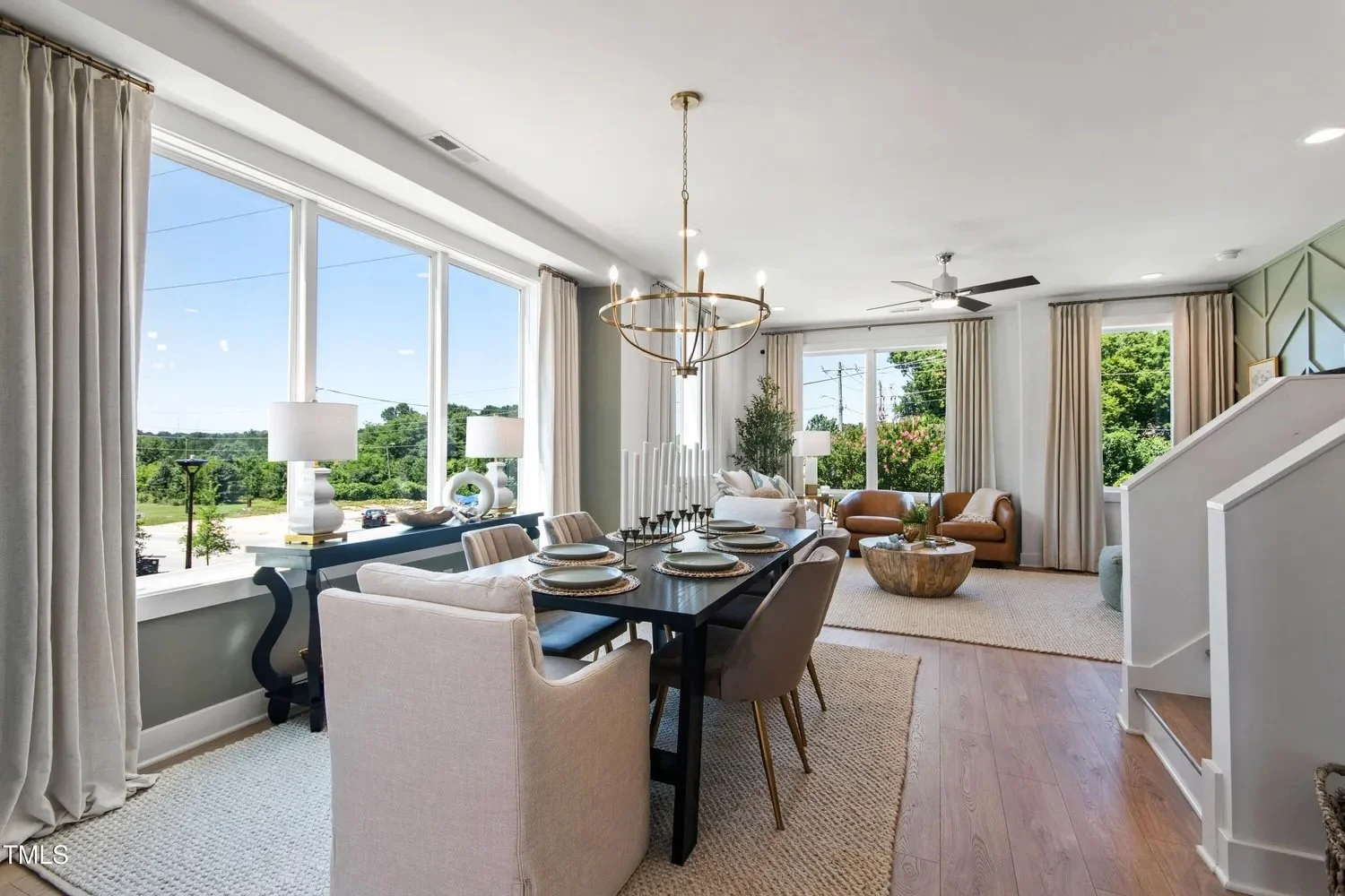 Open-concept living and dining room with large windows, beige curtains, a dark wood dining table with six chairs, a chandelier, a gray sofa, tan armchairs, and a round wooden coffee table. Bright natural light flooding in from outside.