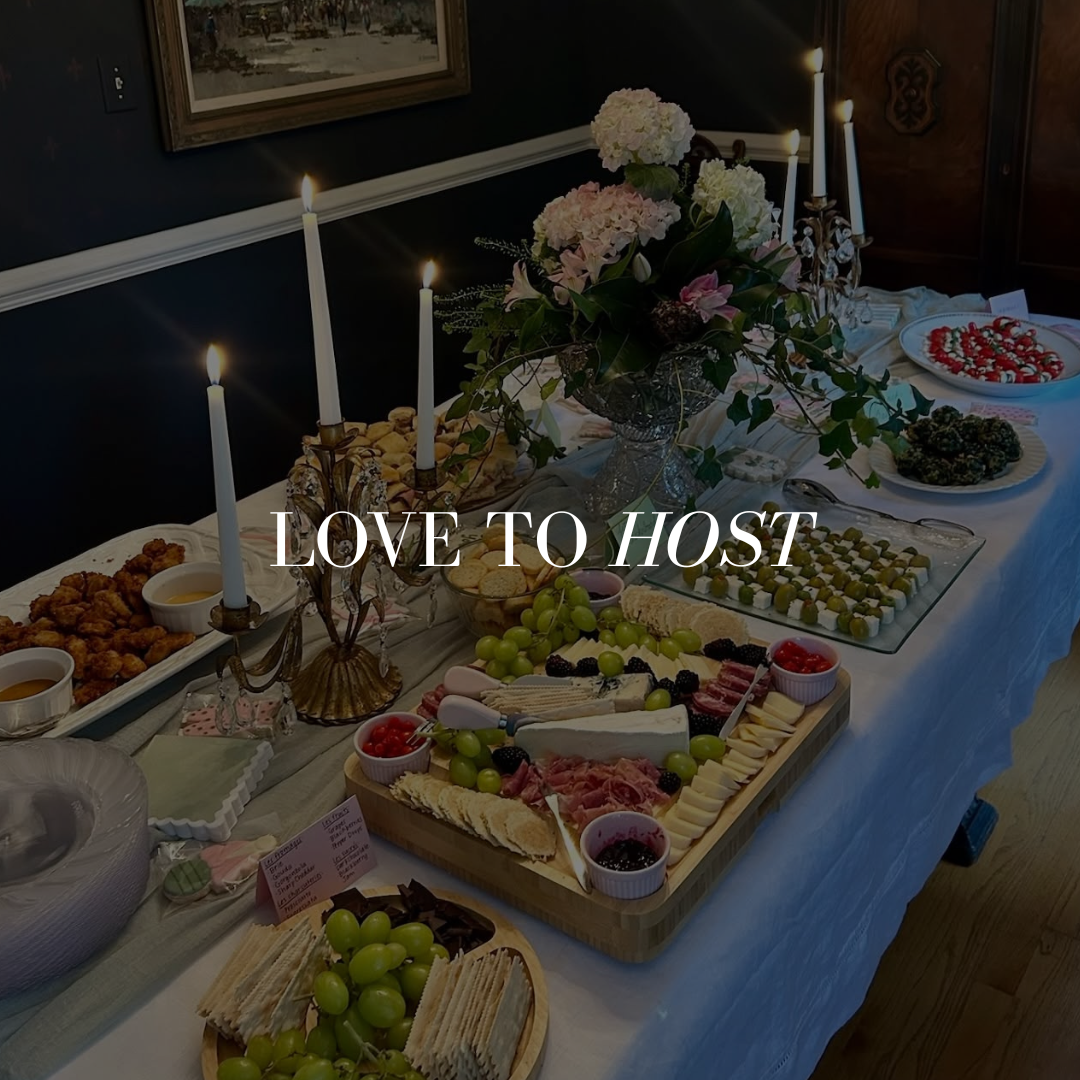 A dinner table with a variety of food including crackers, cheese, grapes, meats, and desserts, decorated with flowers and lit candles. Overlaid text reads "Love to Host."