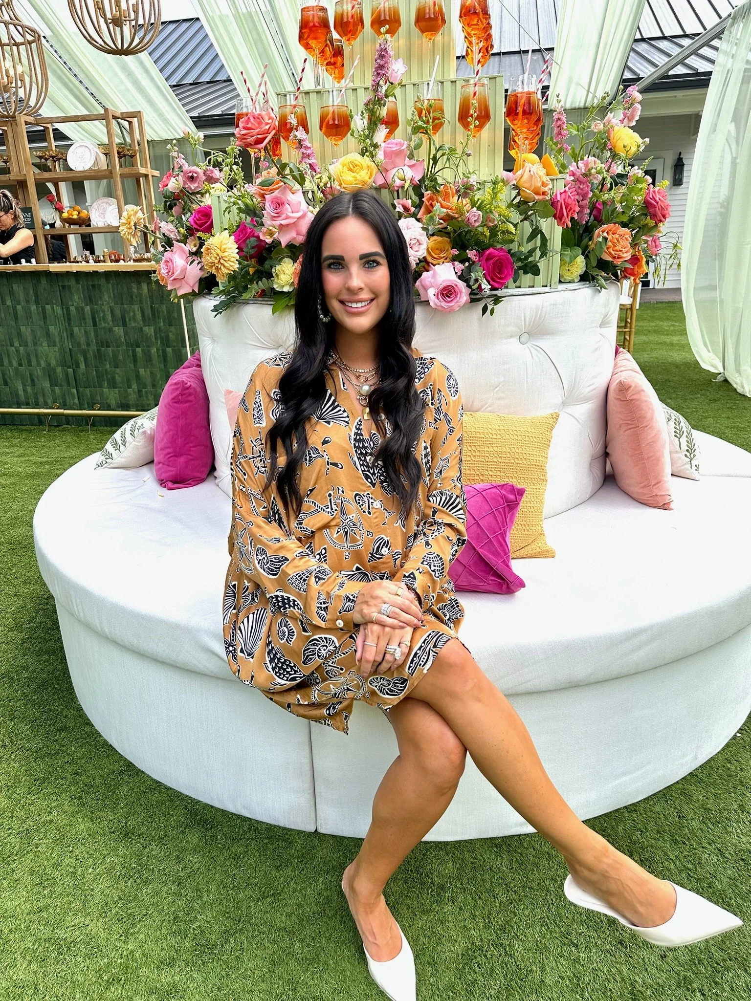 A woman with long black hair and a patterned dress sitting on a large white circular outdoor sofa, in front of a vibrant flower arrangement and hanging glasses with drinks at an outdoor event.