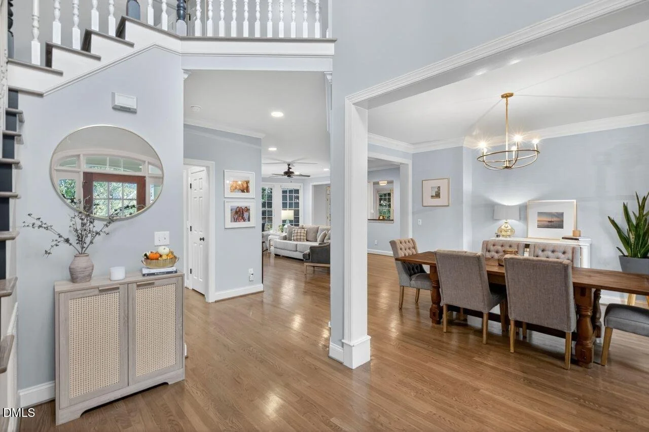 A spacious open-concept living and dining area with hardwood floors, light blue walls, and modern decor. The dining area has six upholstered chairs around a wooden table, a chandelier, and a sideboard with a lamp and framed photo. The living room has a white sofa, a ceiling fan, and large windows letting in natural light.