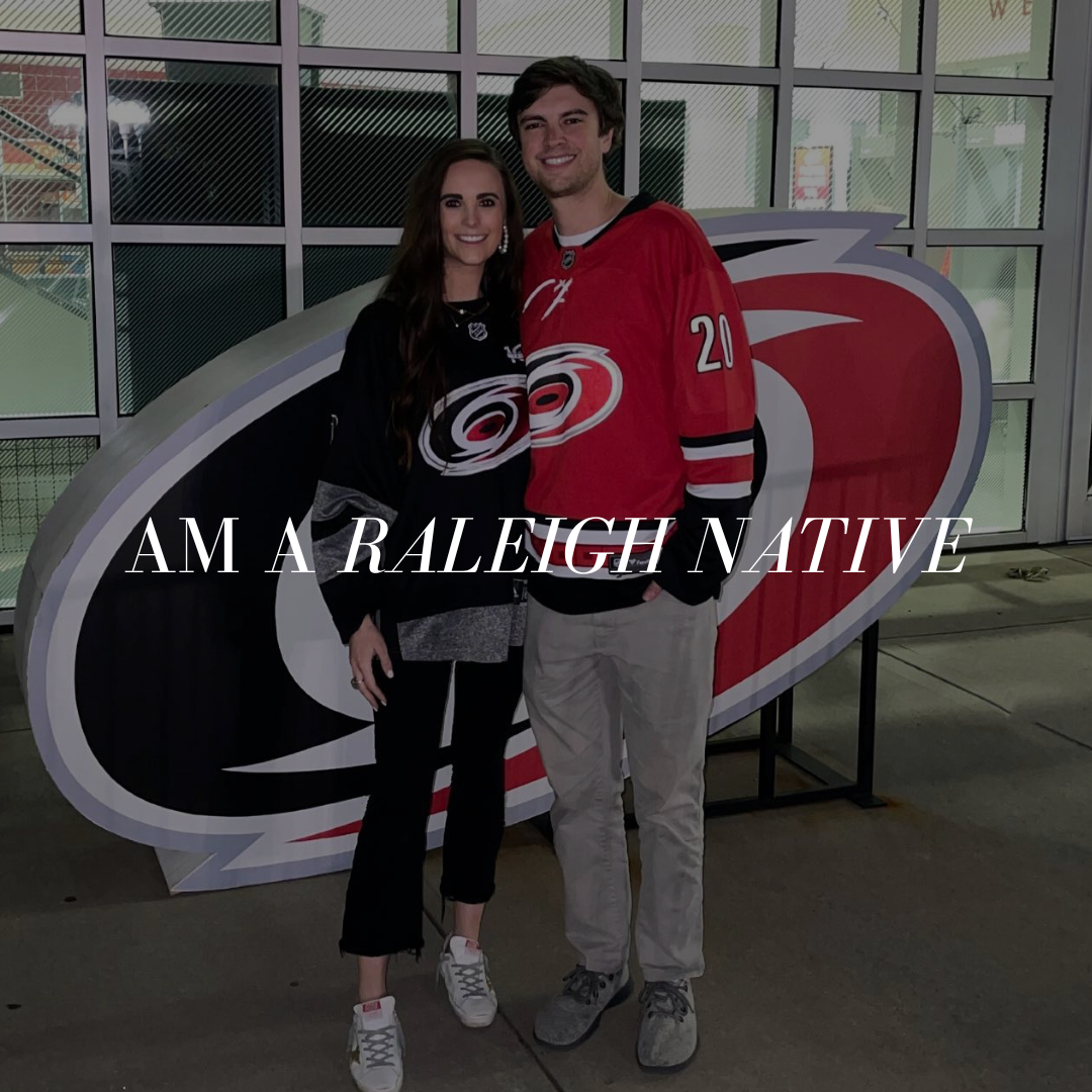 A young couple standing in front of a large team logo at an indoor sports arena. The woman is wearing a black hockey jersey, and the man is wearing a red hockey jersey. The text on the image reads "I am a Raleigh native."