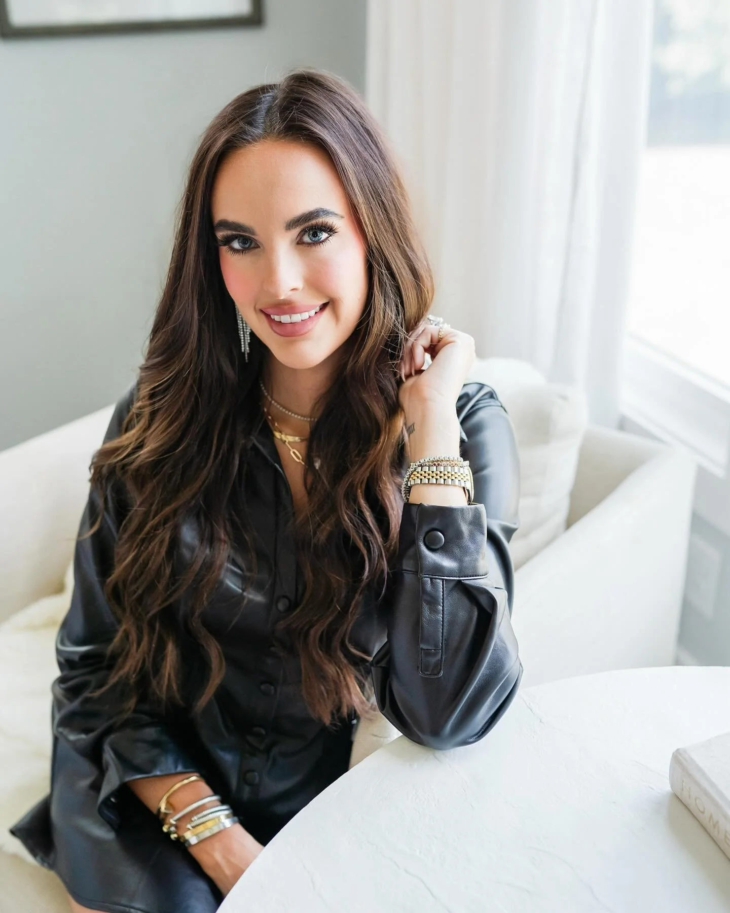 A woman with long brown hair, blue eyes, and makeup sitting on a white sofa near a window with white curtains. She is wearing a black leather jacket, gold jewelry, and smiling at the camera.