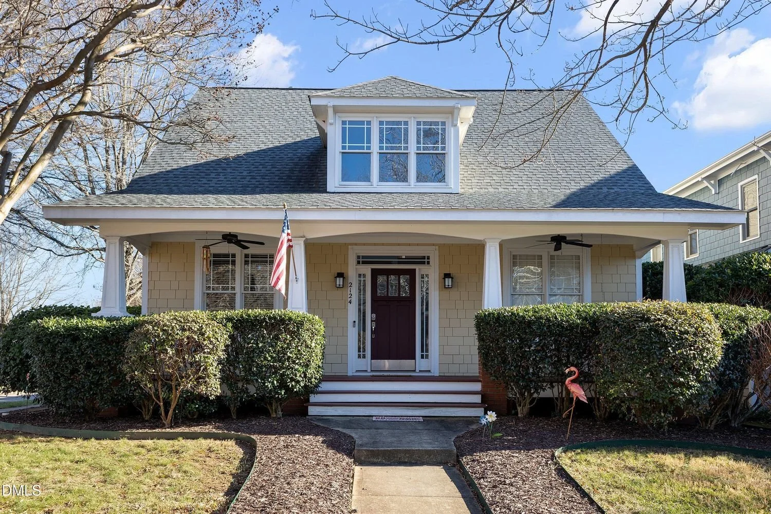 A two-story house with a gabled roof, beige siding, a front porch with white columns, and a dark front door. There are shrubs in front and a butterfly yard ornament on the right side of the walkway.