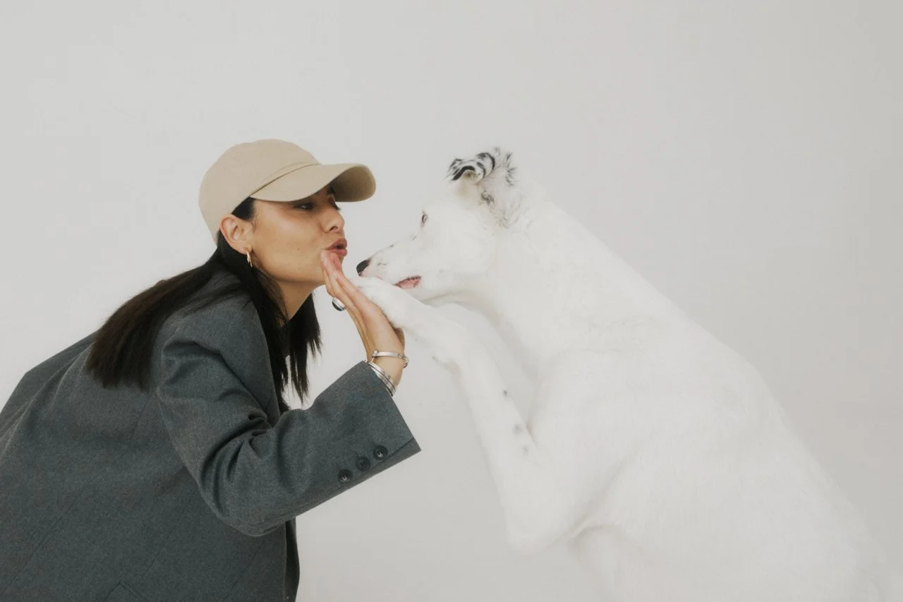 A woman in a beige cap and gray blazer kisses a white dog on the nose.