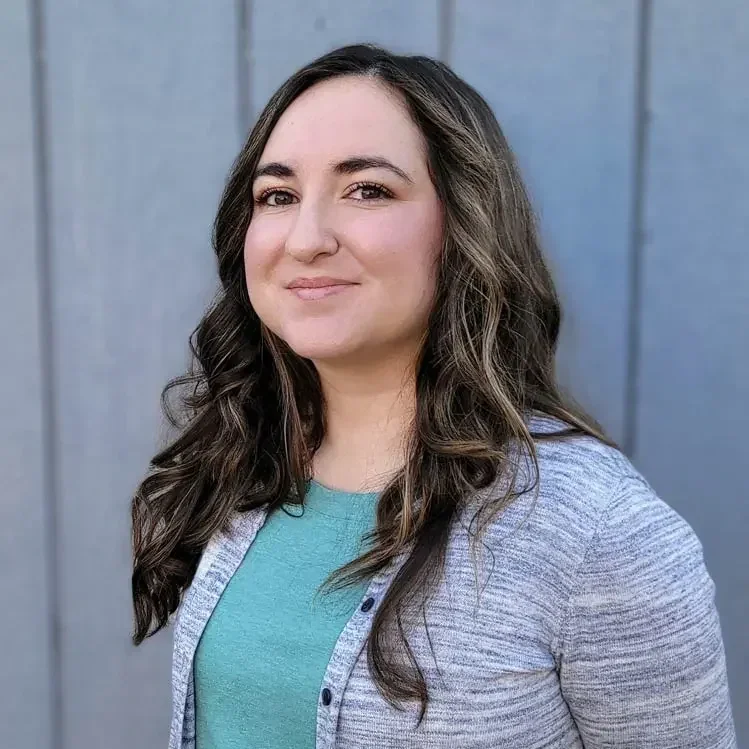 A woman with long dark brown hair smiling and looking at the camera, wearing a gray cardigan over a teal shirt, standing in front of a blue wall.