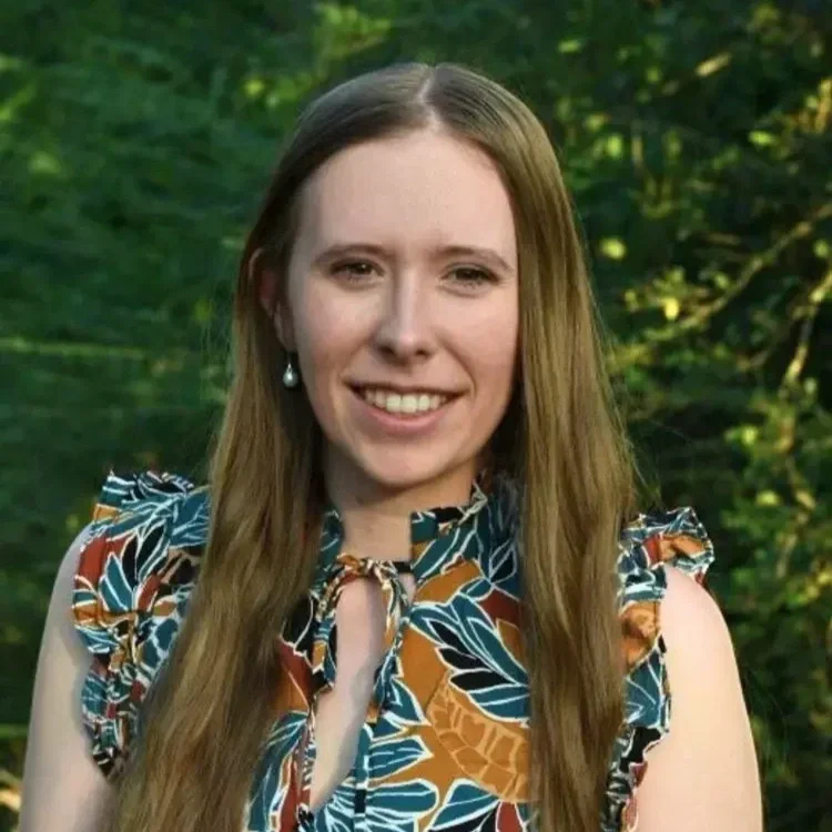 A young woman with long light brown hair, smiling, wearing a patterned sleeveless top with ruffled shoulders and pearl earrings, standing outdoors with green trees in the background.