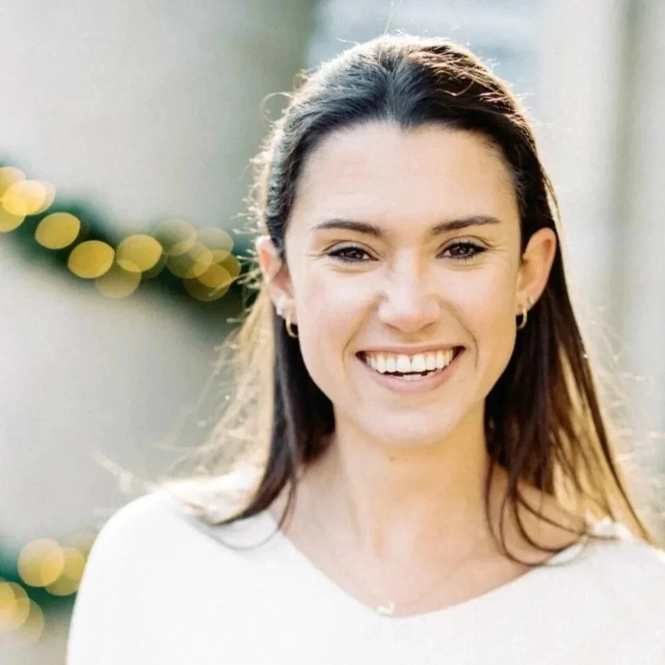 A young woman smiling outdoors with blurred holiday lights in the background.