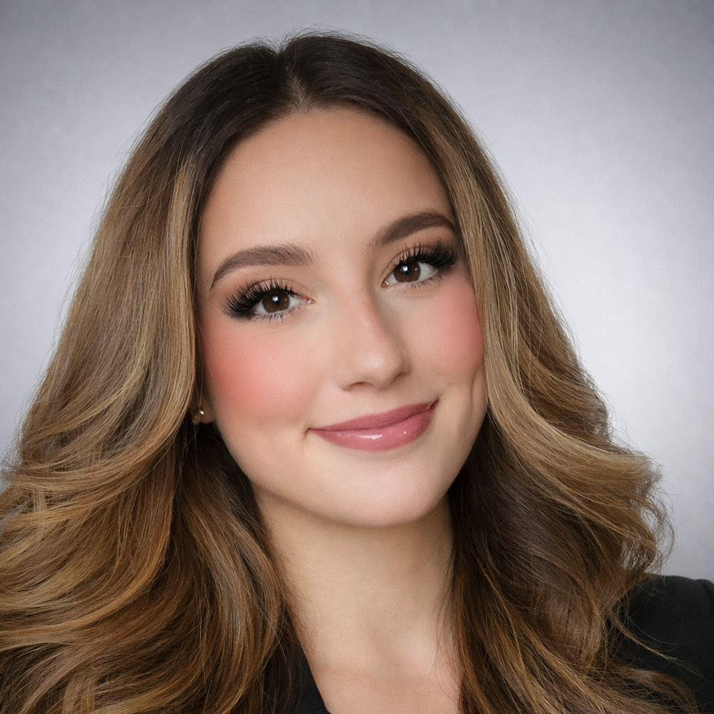 A young woman with long wavy brown hair, wearing makeup and a black top, smiling at the camera against a plain gray background.