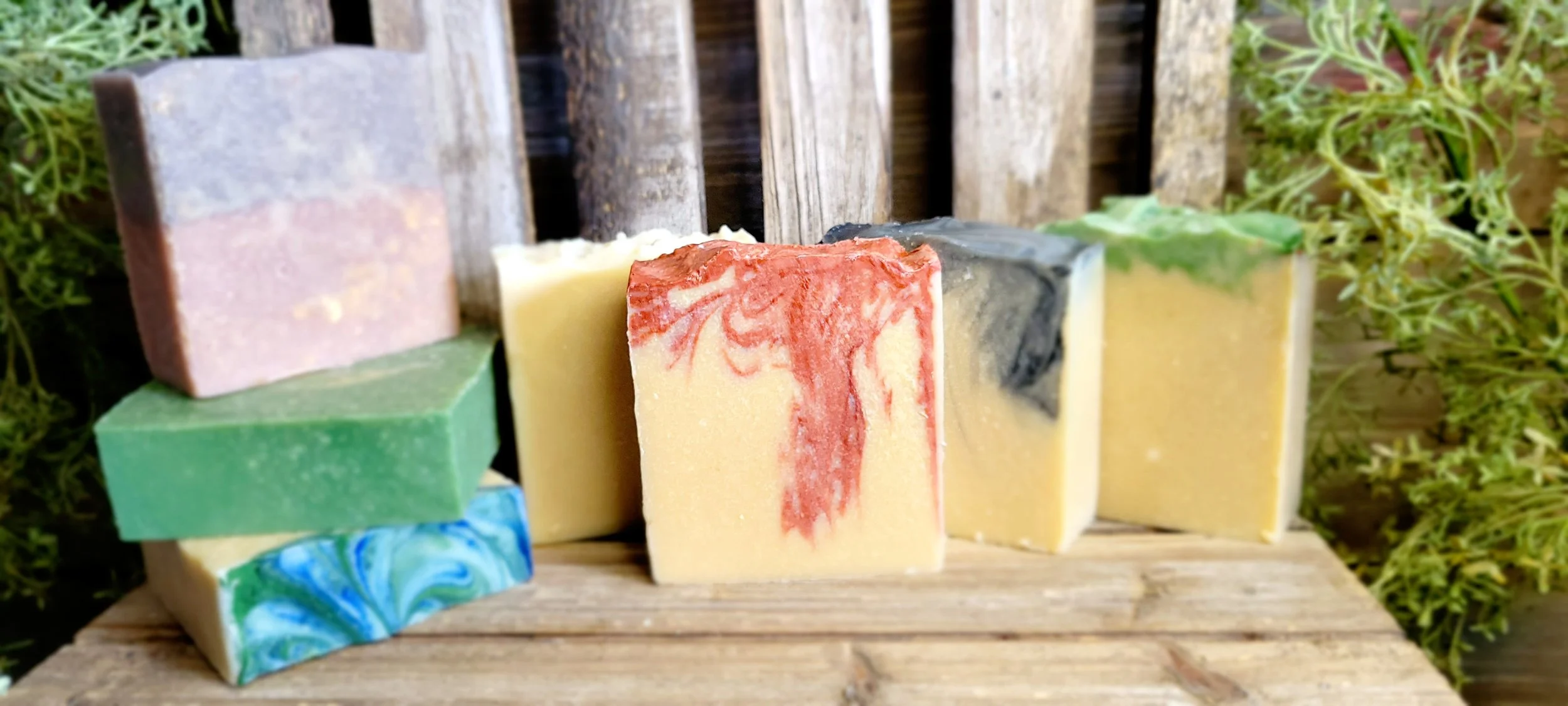 Colorful handmade goat milk soap bars arranged outdoors on a wooden surface with greenery in the background.