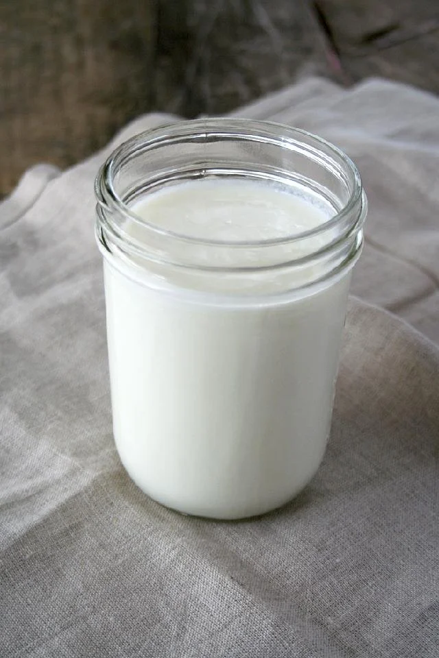 A glass jar filled with goat milk placed on a beige fabric surface.