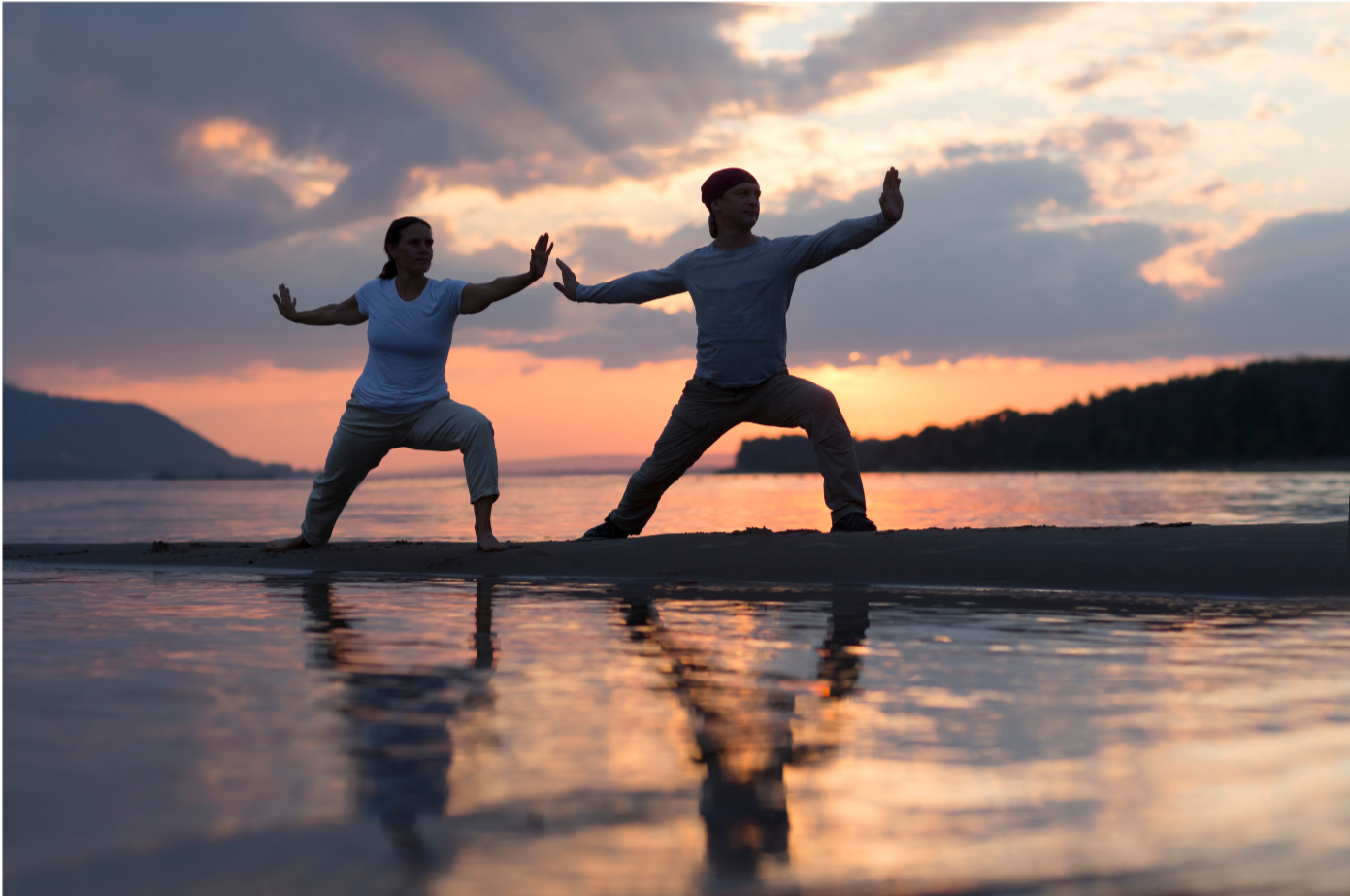 Two people practicing Tai Chi on a beach at sunset with water reflection.