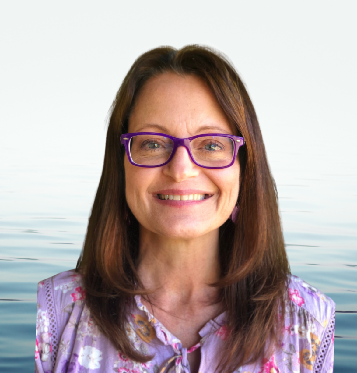 A smiling woman wearing purple glasses and a floral blouse, standing in front of a body of water with ripples and a white sky.