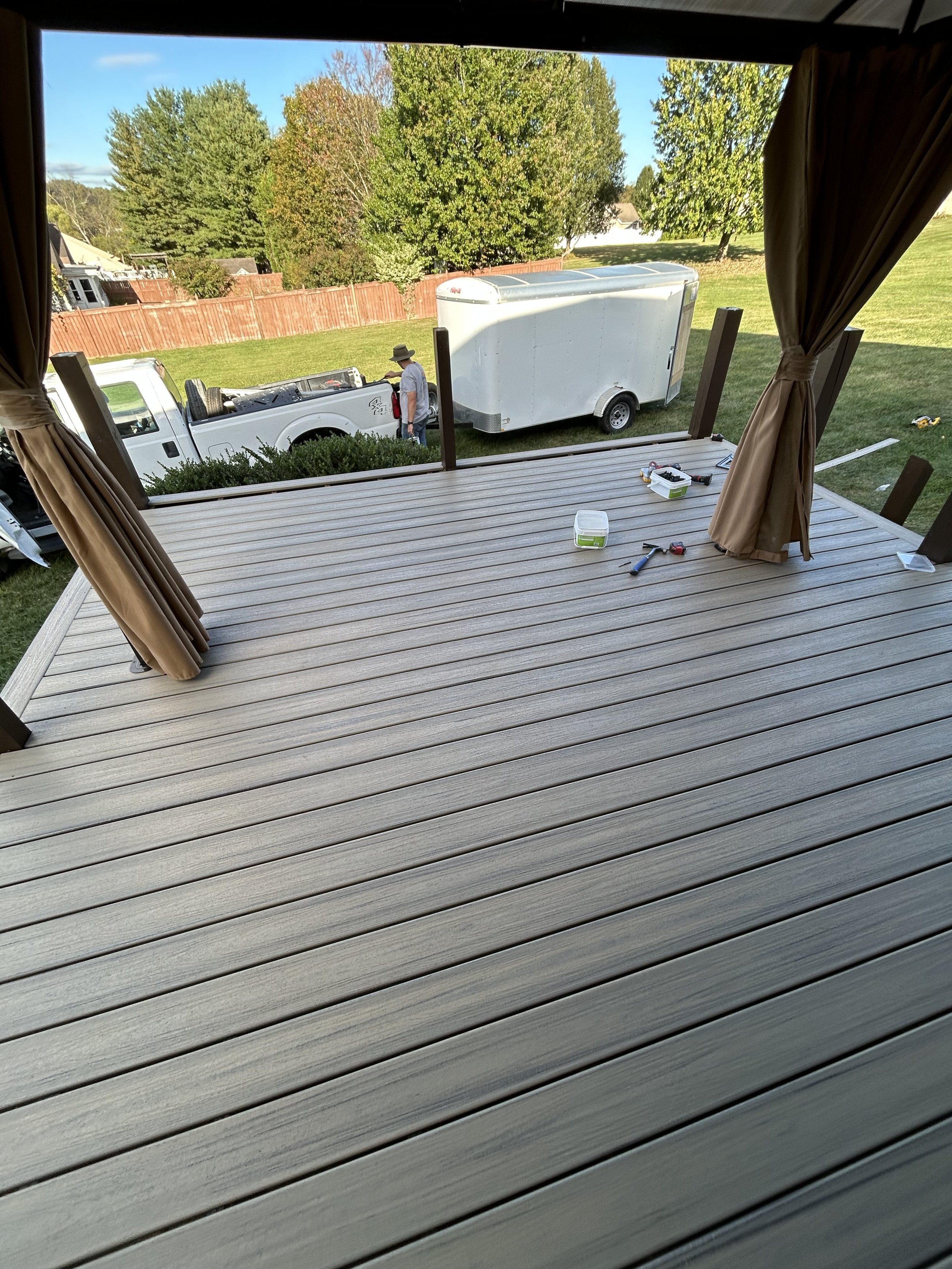 View from a covered porch or deck looking out onto a backyard with a grassy lawn, trees, and a red wooden fence. In the yard, a man wearing a hat is standing near a white pickup truck that is overturned and a white enclosed trailer is parked beside i