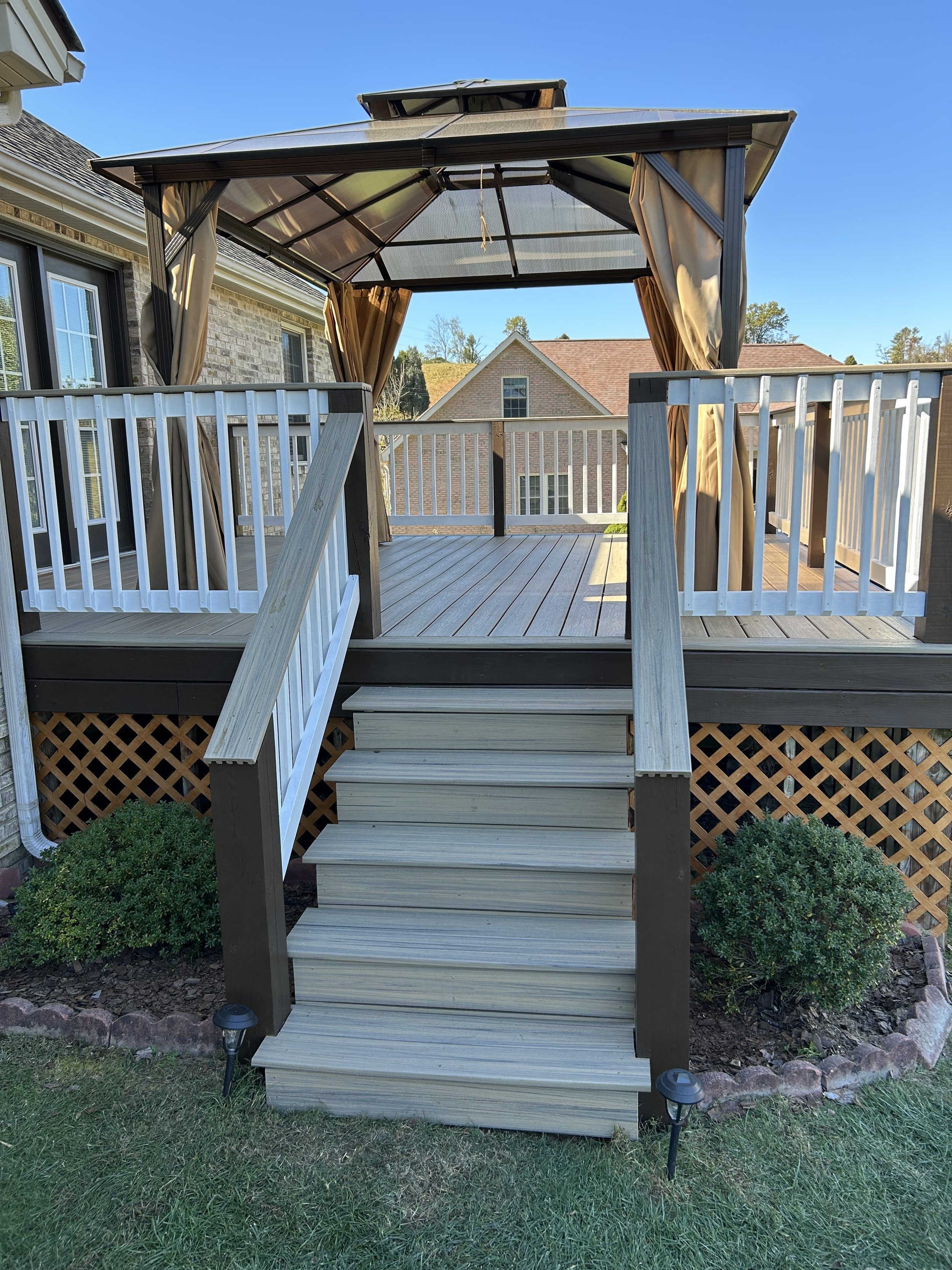 Wooden deck with stairs leading up to a gazebo with curtains, surrounded by small bushes and landscaped garden, in a suburban neighborhood.