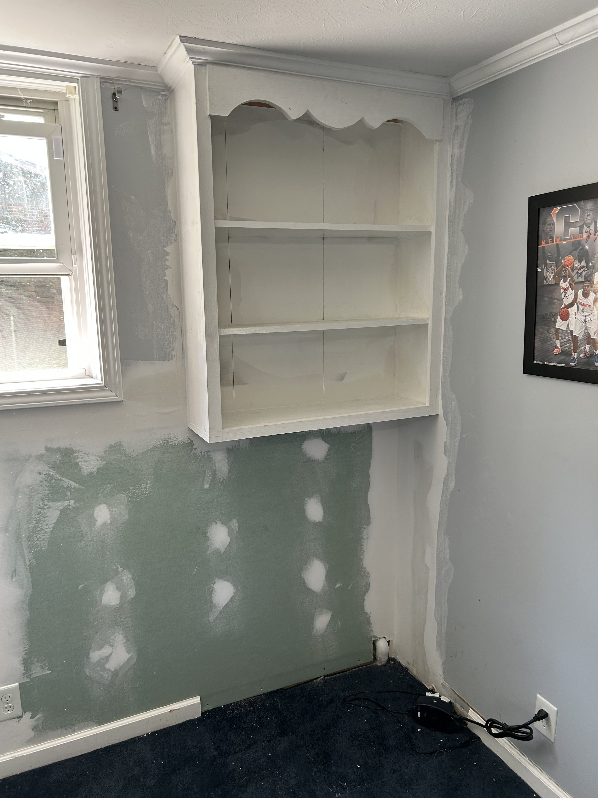A partially painted room with a white cabinet mounted on the wall, a window to the left, and a framed basketball poster to the right. The wall has patches of unpainted drywall with visible joint compound, and an electrical outlet with a black power c