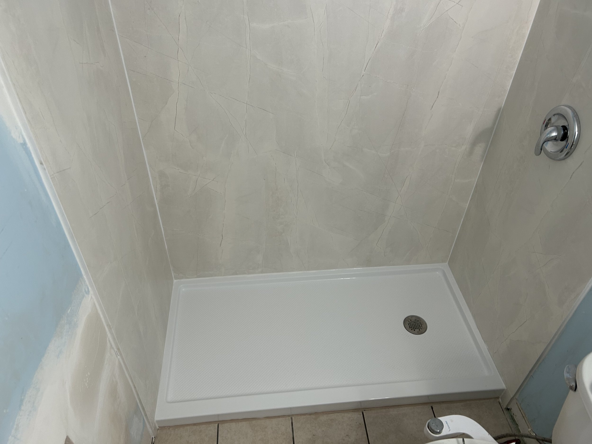 Empty shower stall with a white textured shower base, beige marble-patterned tiles on the walls, a drain in the center of the shower base, and a silver shower faucet handle on the right wall.