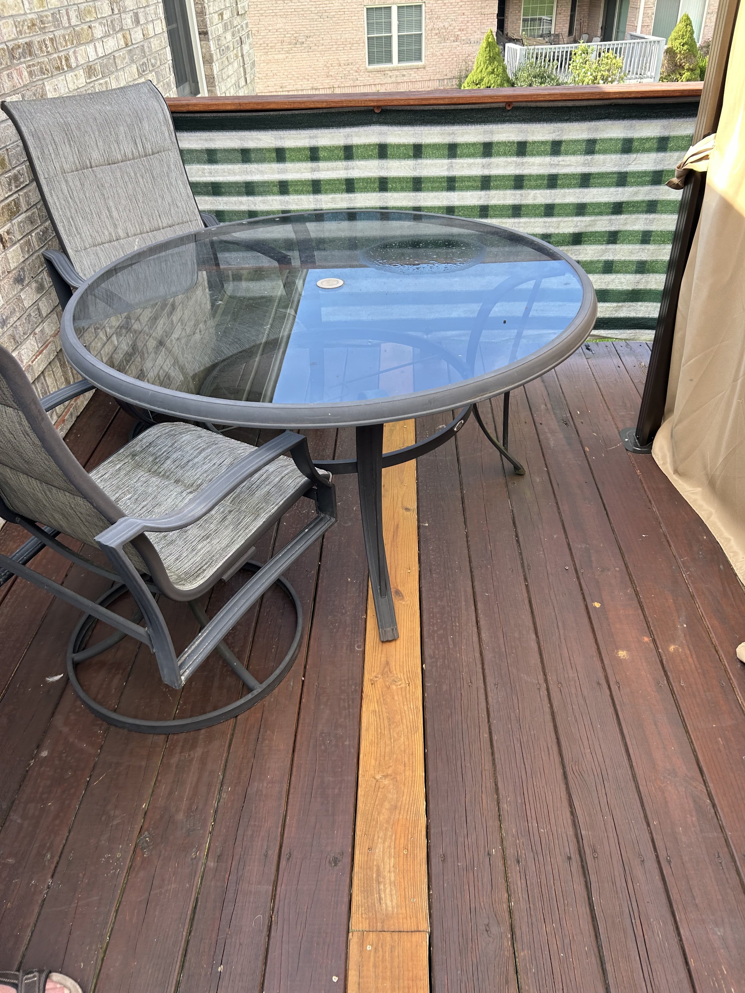 Outdoor balcony with a glass-top table, two chairs, a green checkered fabric privacy screen, and a wooden floor with a narrow wooden strip in the center. Brick building and other balconies are visible in the background.