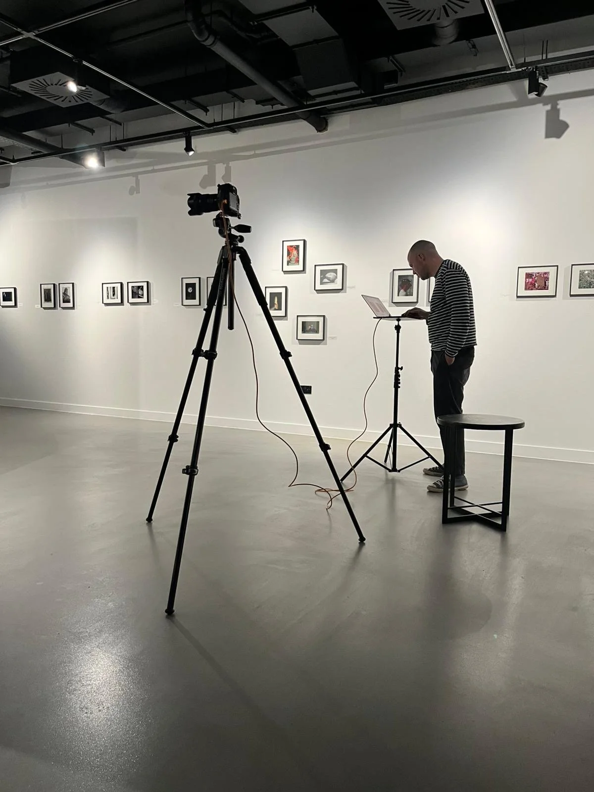 An individual standing in an art gallery, using a laptop music stand, with framed artwork on the wall behind. A camera on a tripod is set up to record or photograph the scene.