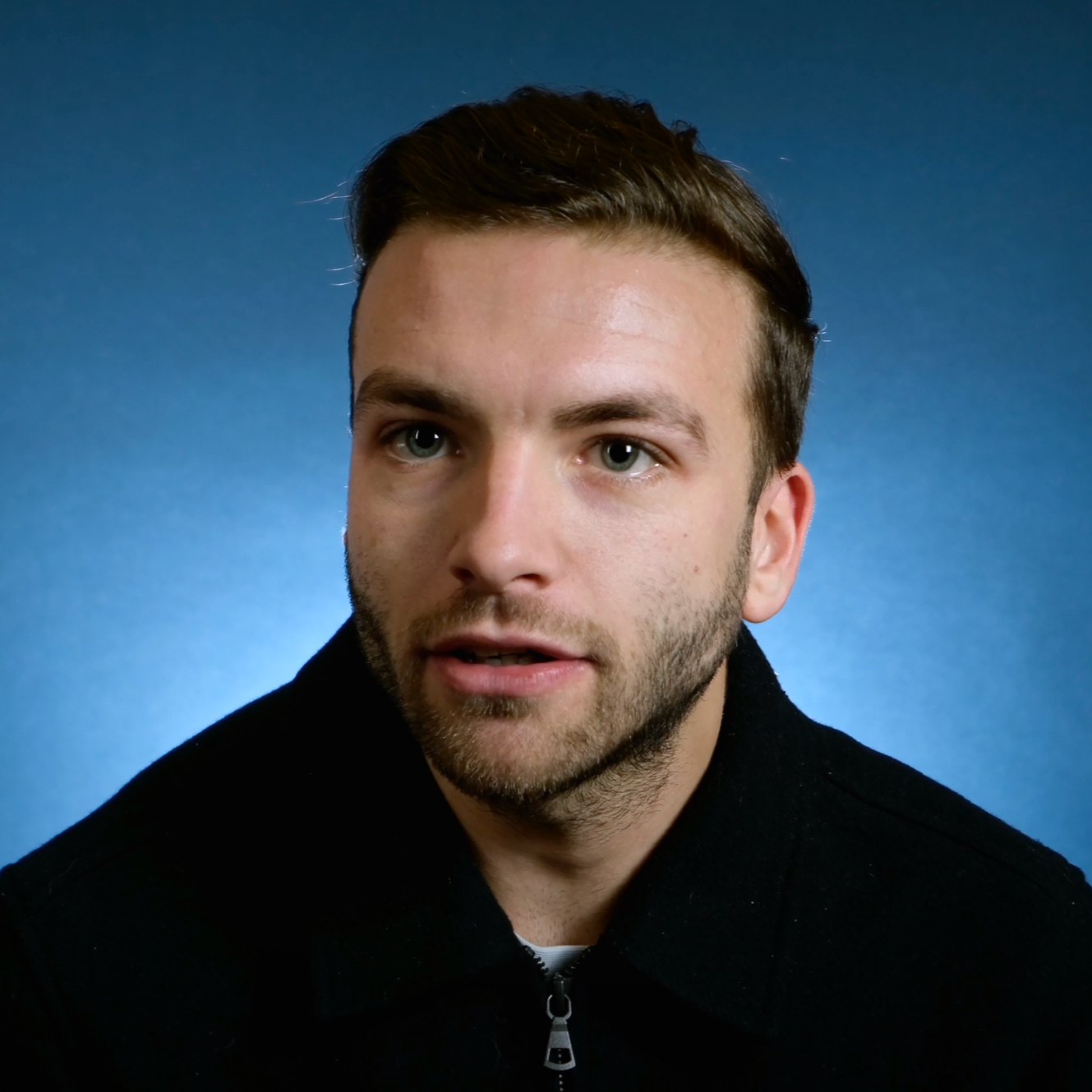 Portrait of a young man with brown hair, blue eyes, and a short beard, wearing a black zippered jacket against a blue gradient background.