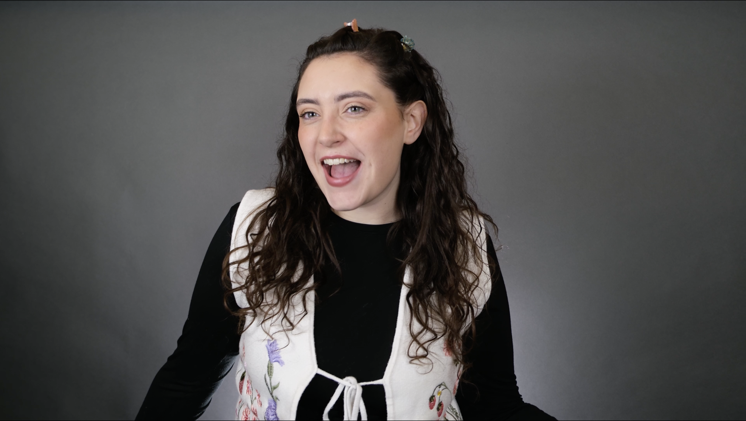 A young woman with long curly brown hair, wearing a black top and a white embroidered vest, smiling and looking at the camera against a plain gray background.