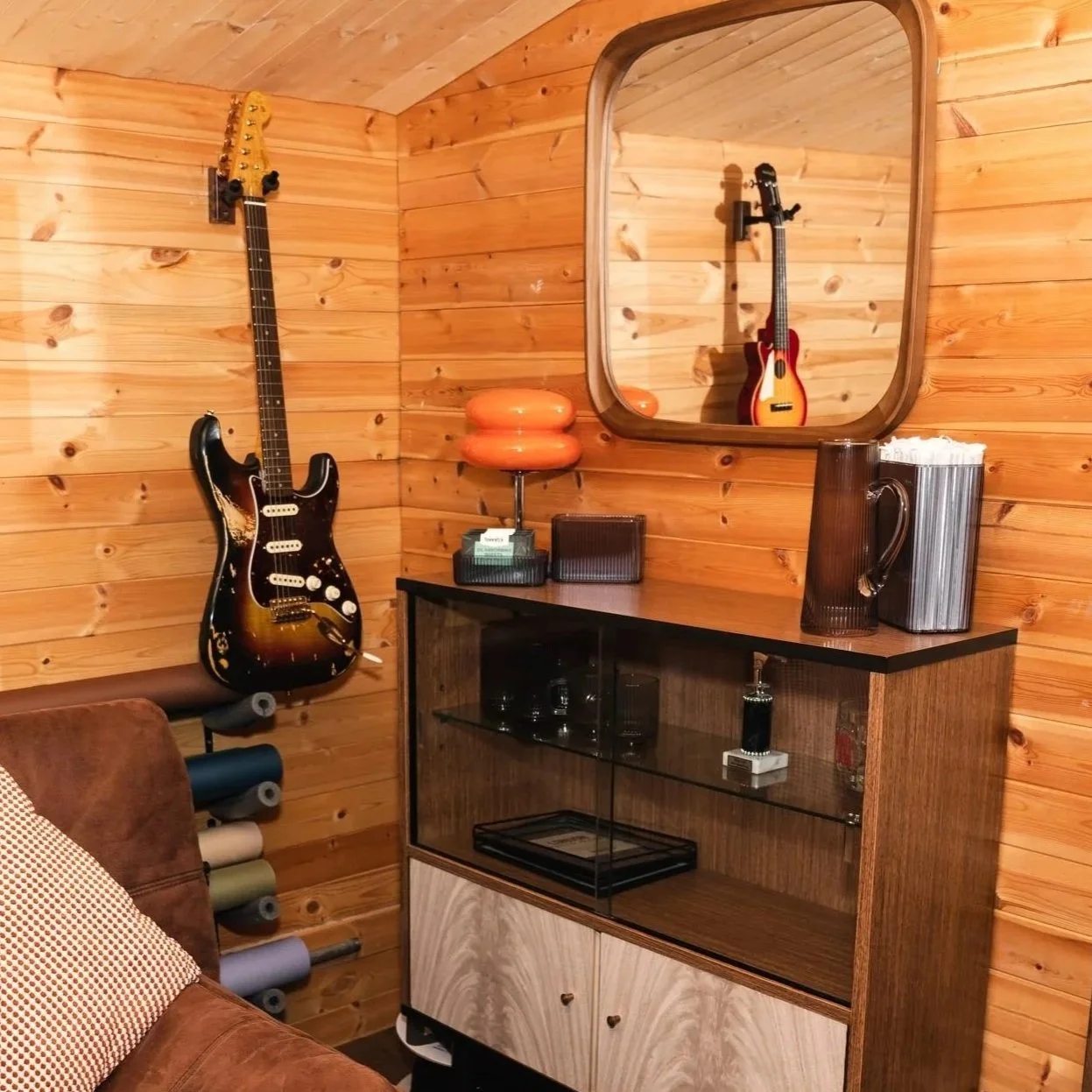 A corner with wood-paneled walls and ceiling, featuring an electric guitar hanging on the wall, a large mirror reflecting the guitar, a wooden cabinet with glass doors displaying glassware, a tall striped pitcher, a small brown pitcher, a vintage orange lamp, a box of tissues, and a brown sofa with a patterned cushion.